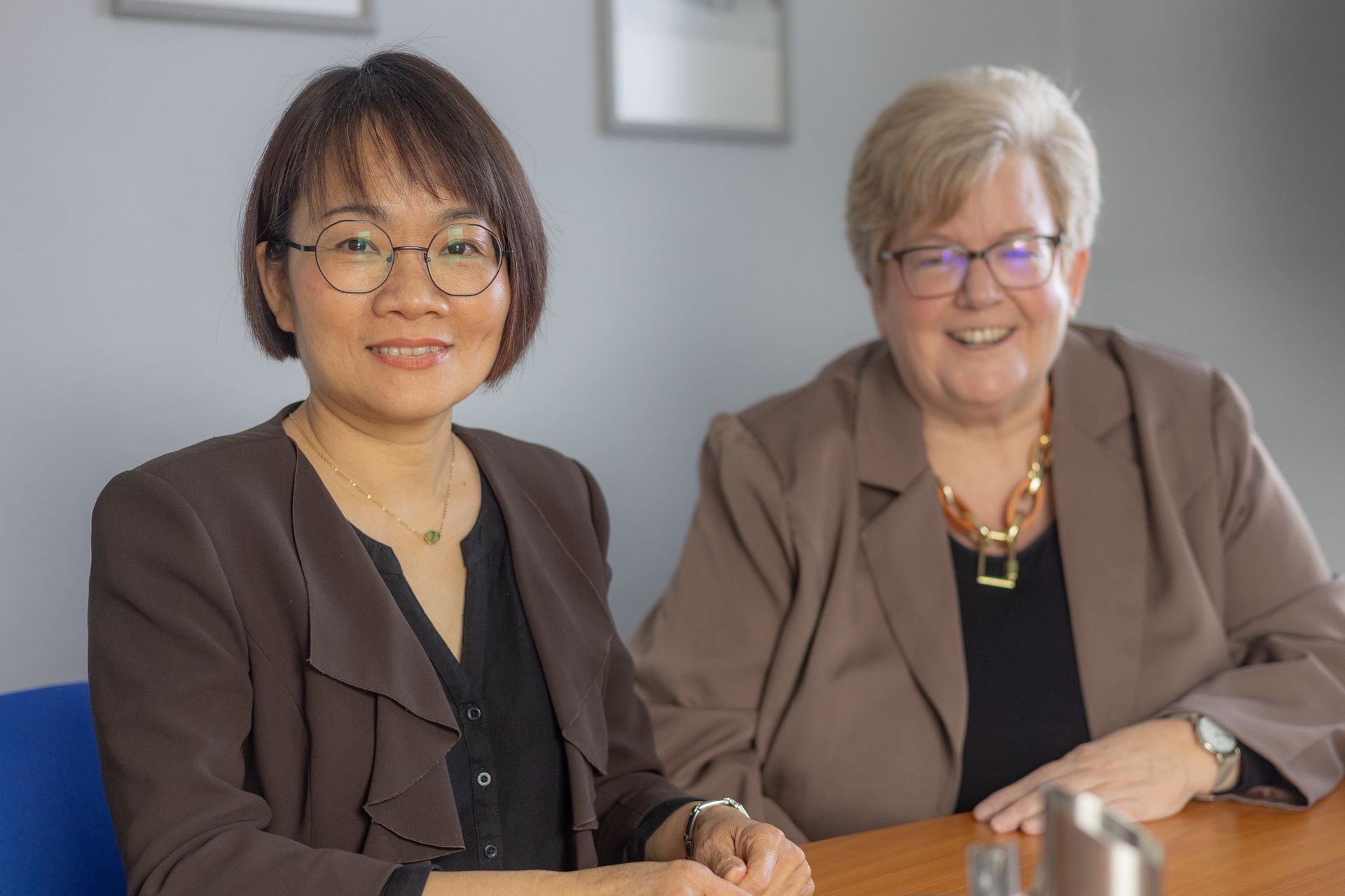Zwei Frauen sitzen an einem Tisch und lächeln in die Kamera. Sie tragen Businesskleidung und befinden sich in einem professionellen Umfeld.