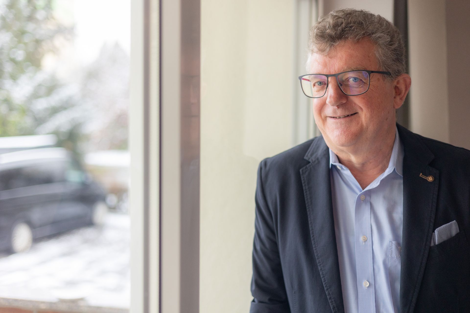 Ein Mann mit Brille lächelt und schaut aus dem Fenster. Er trägt ein blaues Hemd und einen Blazer, natürliches Licht.