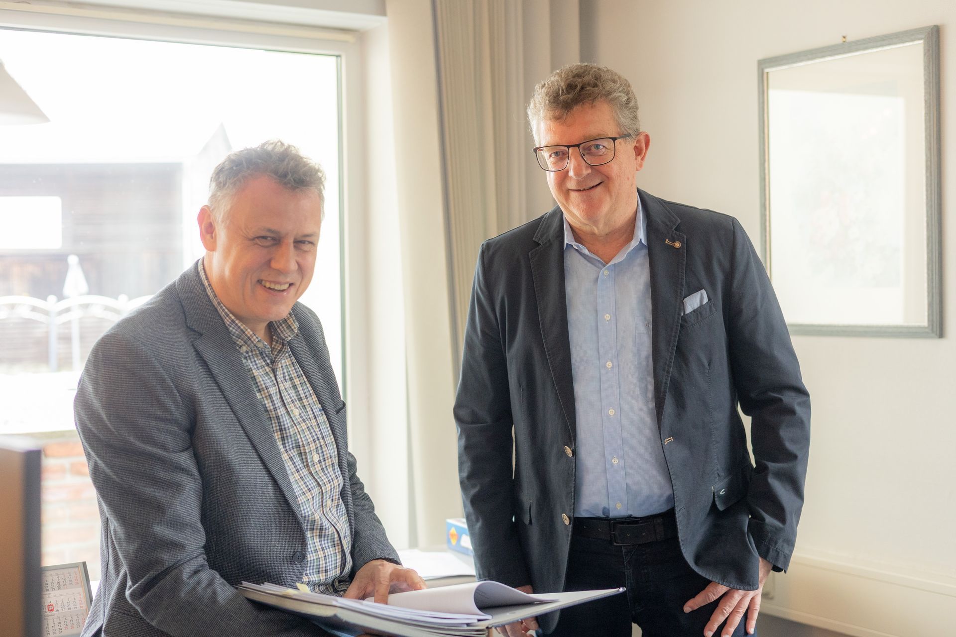 Zwei lächelnde Männer in Geschäftskleidung, einer sitzt und betrachtet Dokumente, der andere steht in der Nähe eines Fensters.