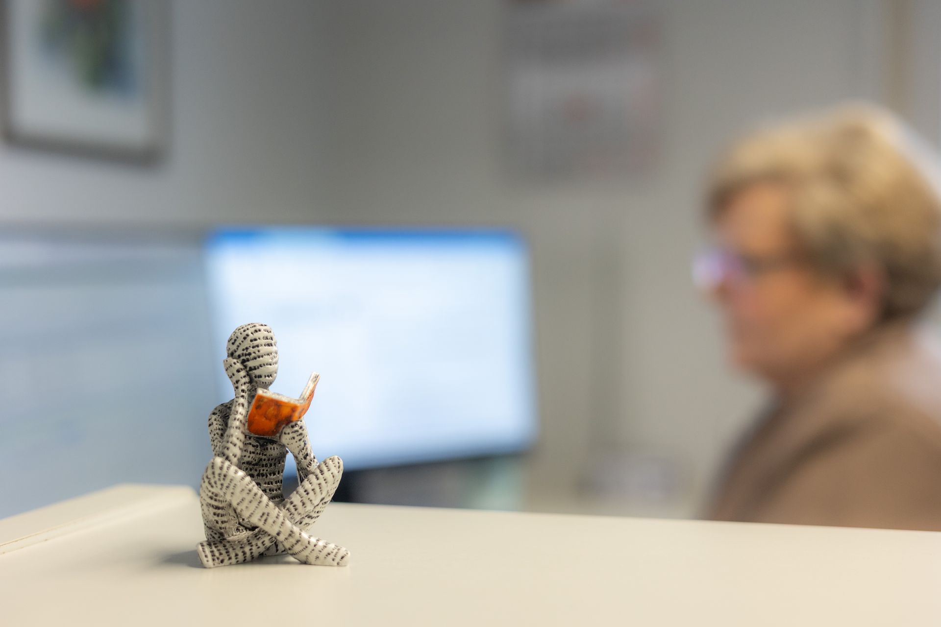 Eine kleine Figur aus Papier liest ein Buch, im verschwommenen Hintergrund sind eine Person und ein Computer zu sehen.
