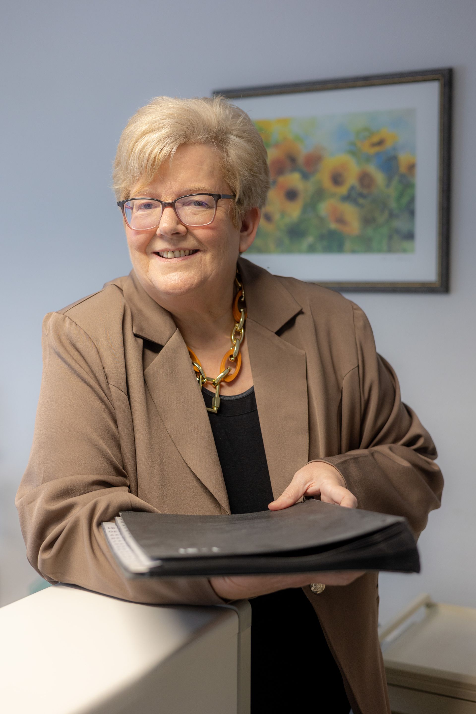 Eine Frau mit Brille und brauner Jacke lächelt und hält Akten in der Hand vor einer hellen Wand mit einem gerahmten Blumendruck.