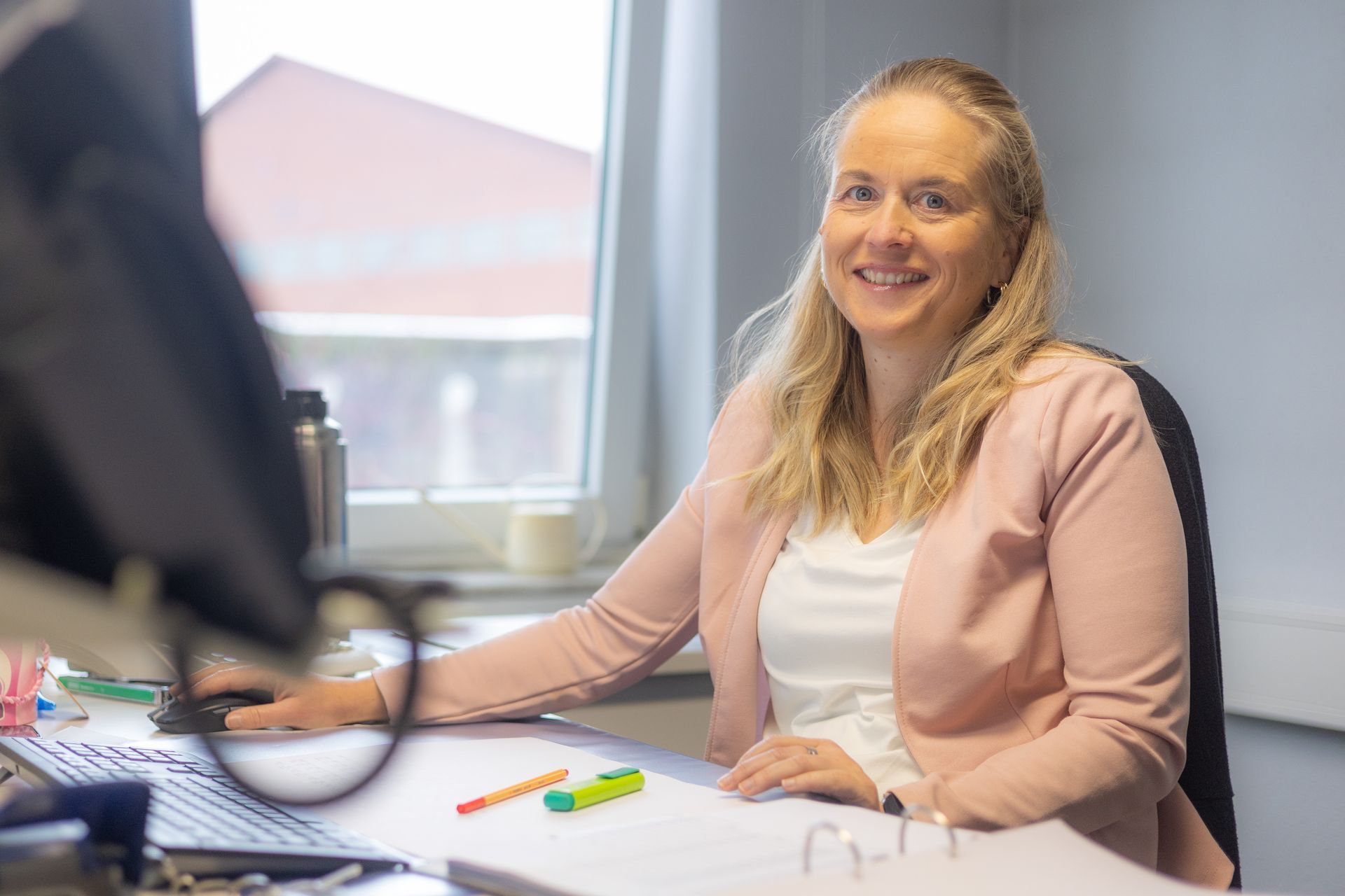 Frau lächelt bei der Arbeit an einem Schreibtisch, hellrosa Blazer, im Hintergrund ein Fenster.