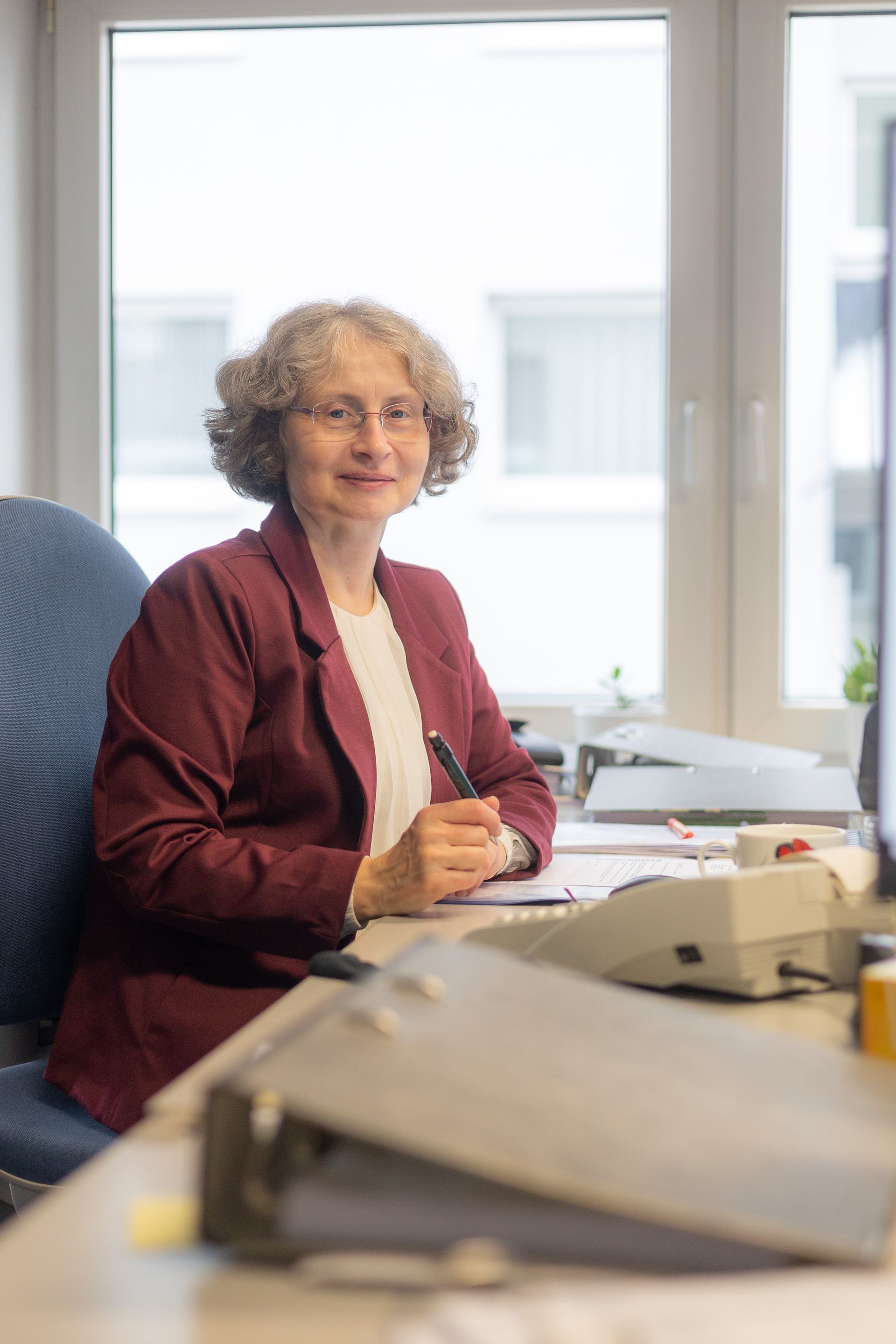 Eine Frau in einem bordeauxroten Blazer lächelt, sie sitzt an einem Schreibtisch in der Nähe eines Fensters und hält einen Stift in der Hand.