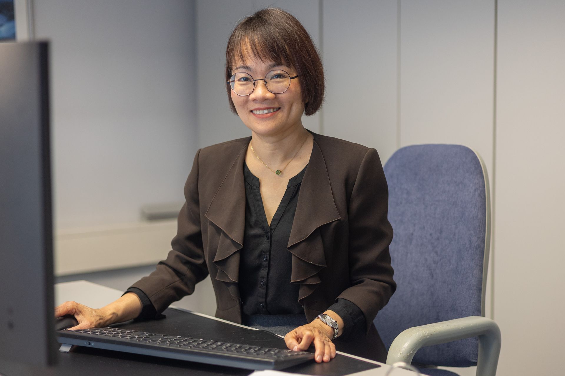 Frau sitzt lächelnd an einem Schreibtisch, trägt eine Brille und einen dunklen Blazer; tippt auf einer Tastatur in einer Büroumgebung.