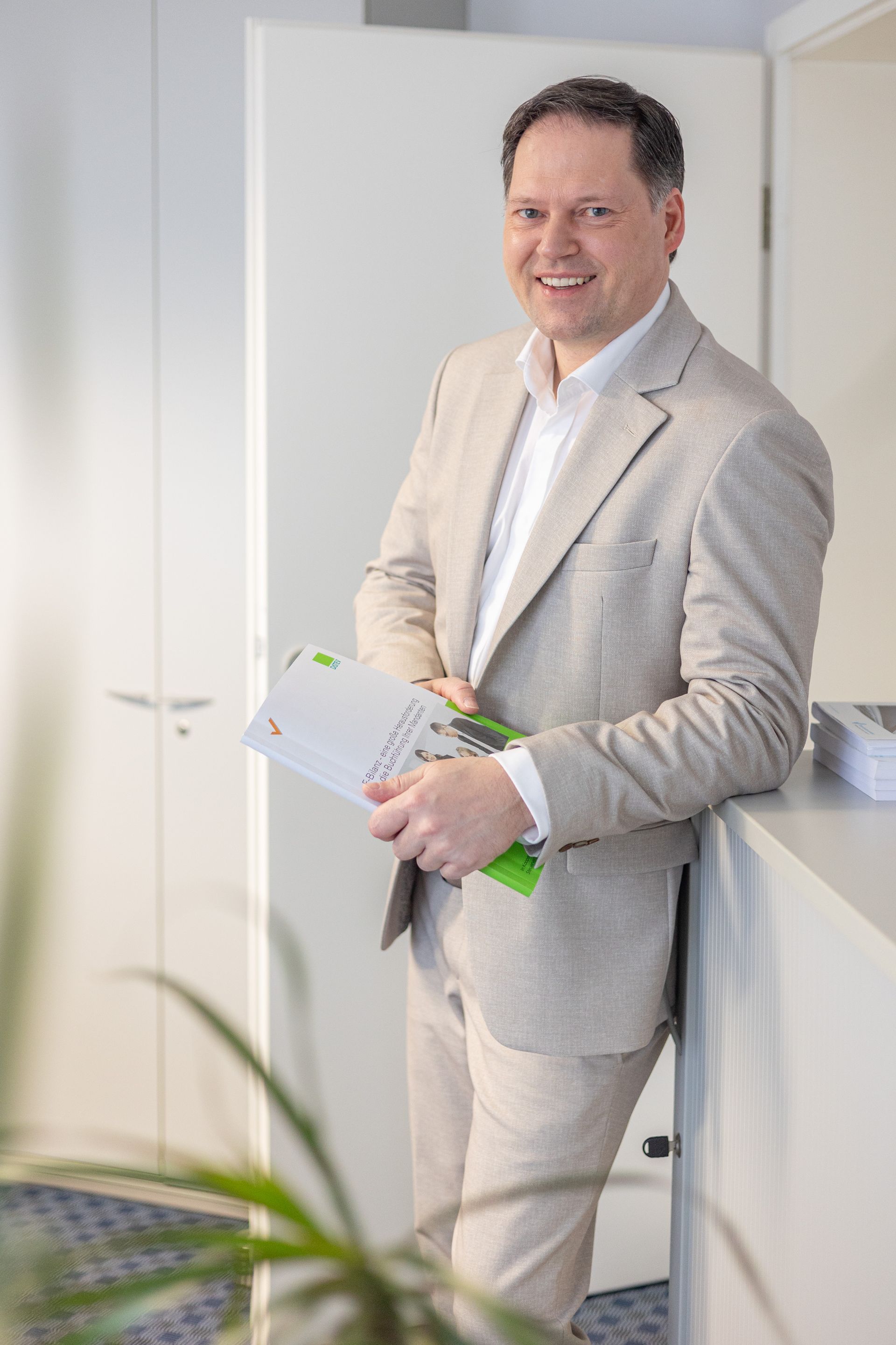 Ein Mann in einem hellen Anzug lächelt, hält eine Broschüre in der Hand und lehnt an einem weißen Tresen in einem Büro.