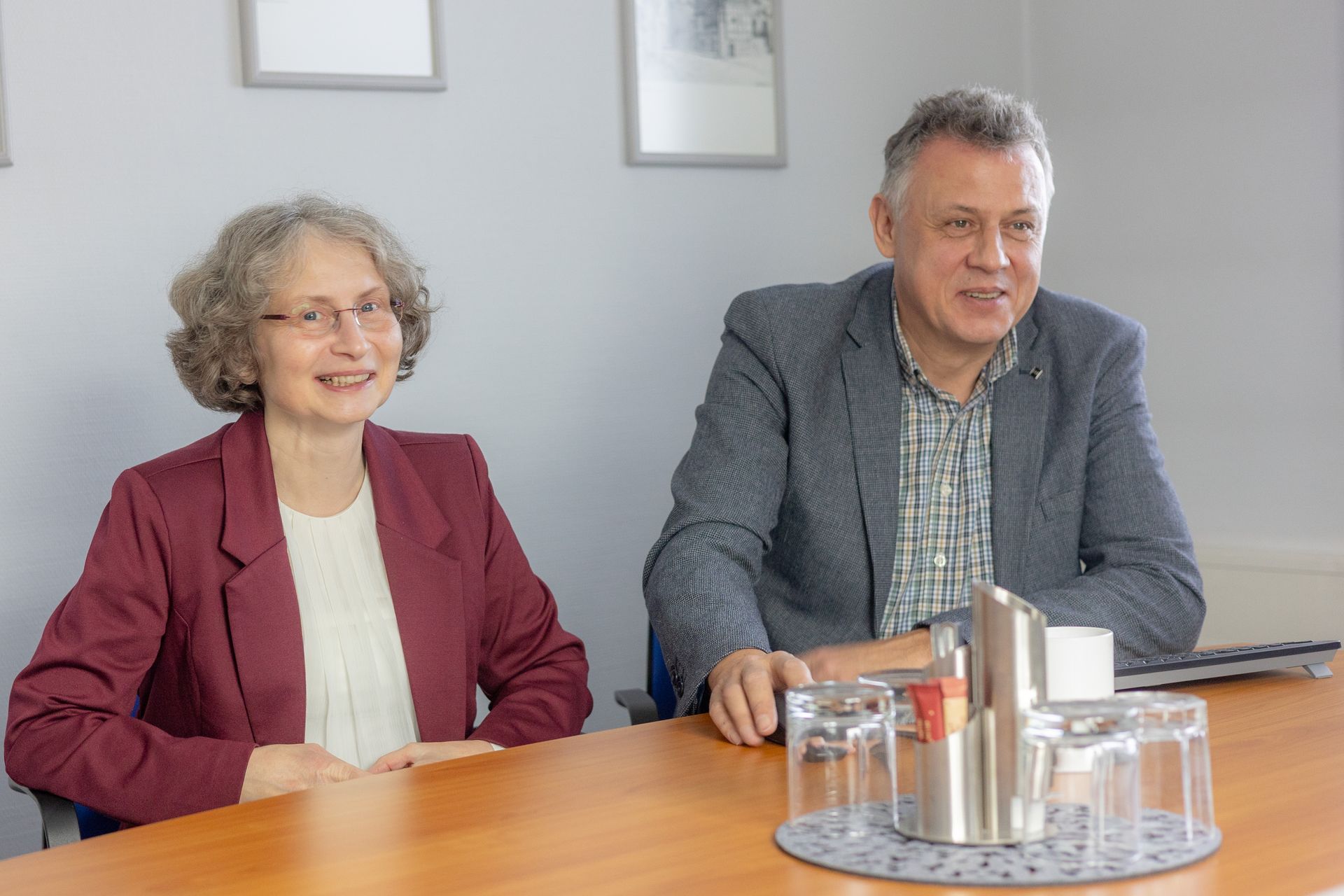 Zwei Personen sitzen lächelnd an einem Tisch. Die Frau trägt einen roten Blazer. Der Mann trägt eine blaue Jacke.