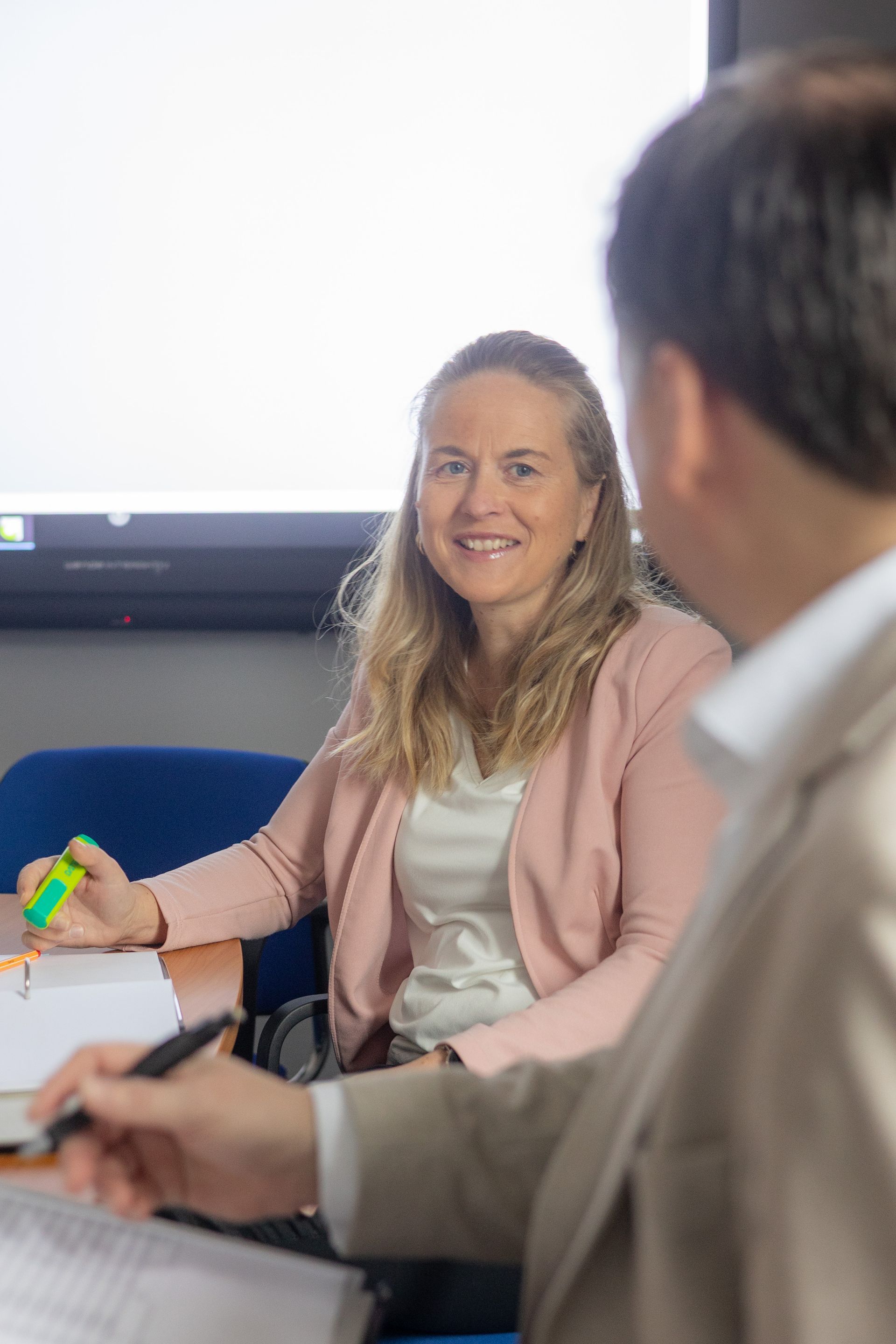 Frau lächelt, sitzt an einem Tisch und blickt zu einer Person außerhalb des Bildausschnitts; sie trägt eine rosa Strickjacke.