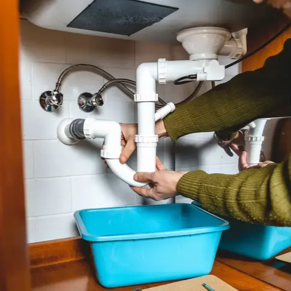 Fontanero arreglando tuberías del fregadero, con las manos sobre tuberías de PVC blanco, lavabo azul debajo.