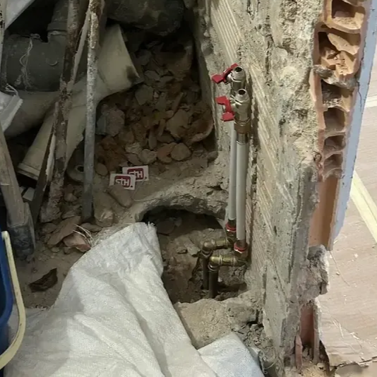 Tuberías con válvulas rojas instaladas en un muro parcialmente demolido. Escombros y herramientas rodean el área de trabajo.