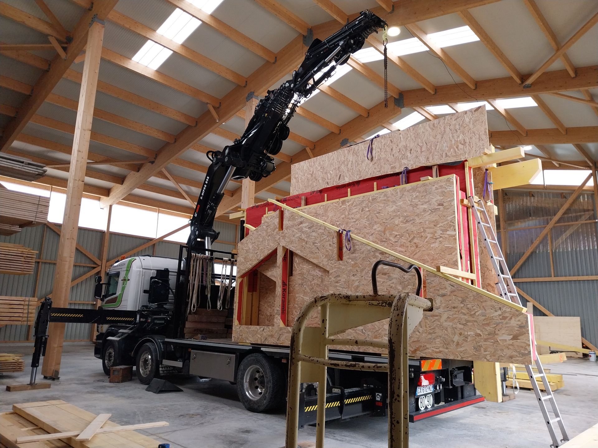 Une grue soulève une grande structure en bois à l'intérieur d'un atelier.