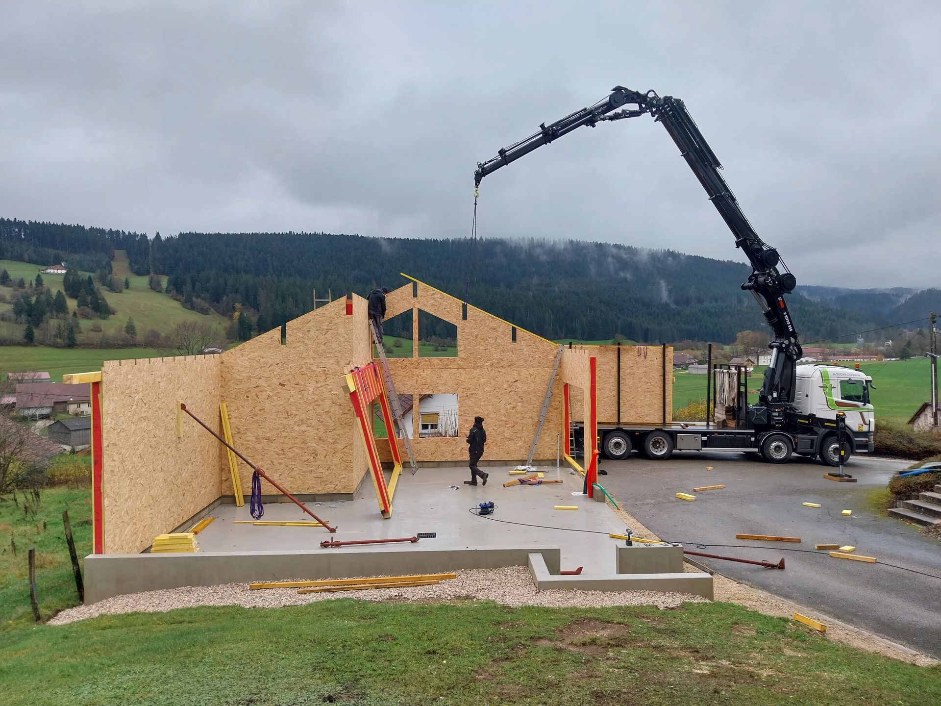 Chantier de construction : une grue soulève un panneau mural en bois pour le poser sur les fondations d'un bâtiment, sous le regard d'un ouvrier.
