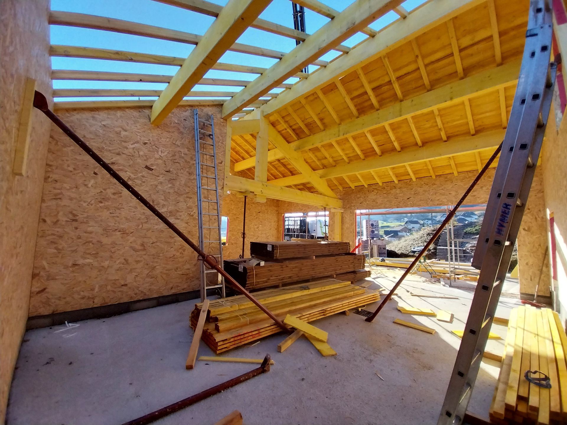 Intérieur d'un bâtiment en construction, charpente en bois, toit ouvert, bois d'œuvre, échelles, supports, ciel dégagé.