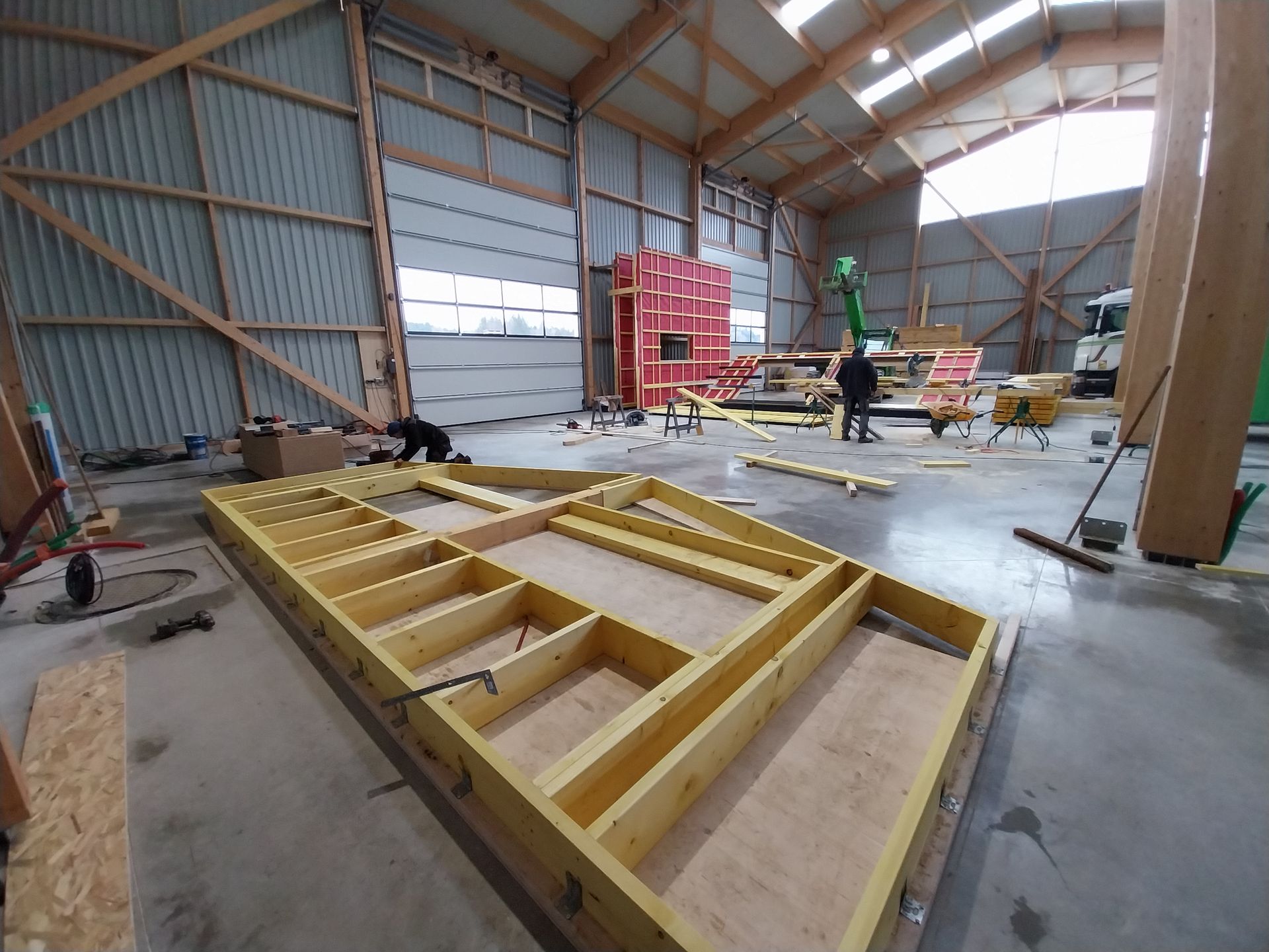 Intérieur d'un chantier de construction : ossature bois au sol, ouvriers, ambiance d'entrepôt.