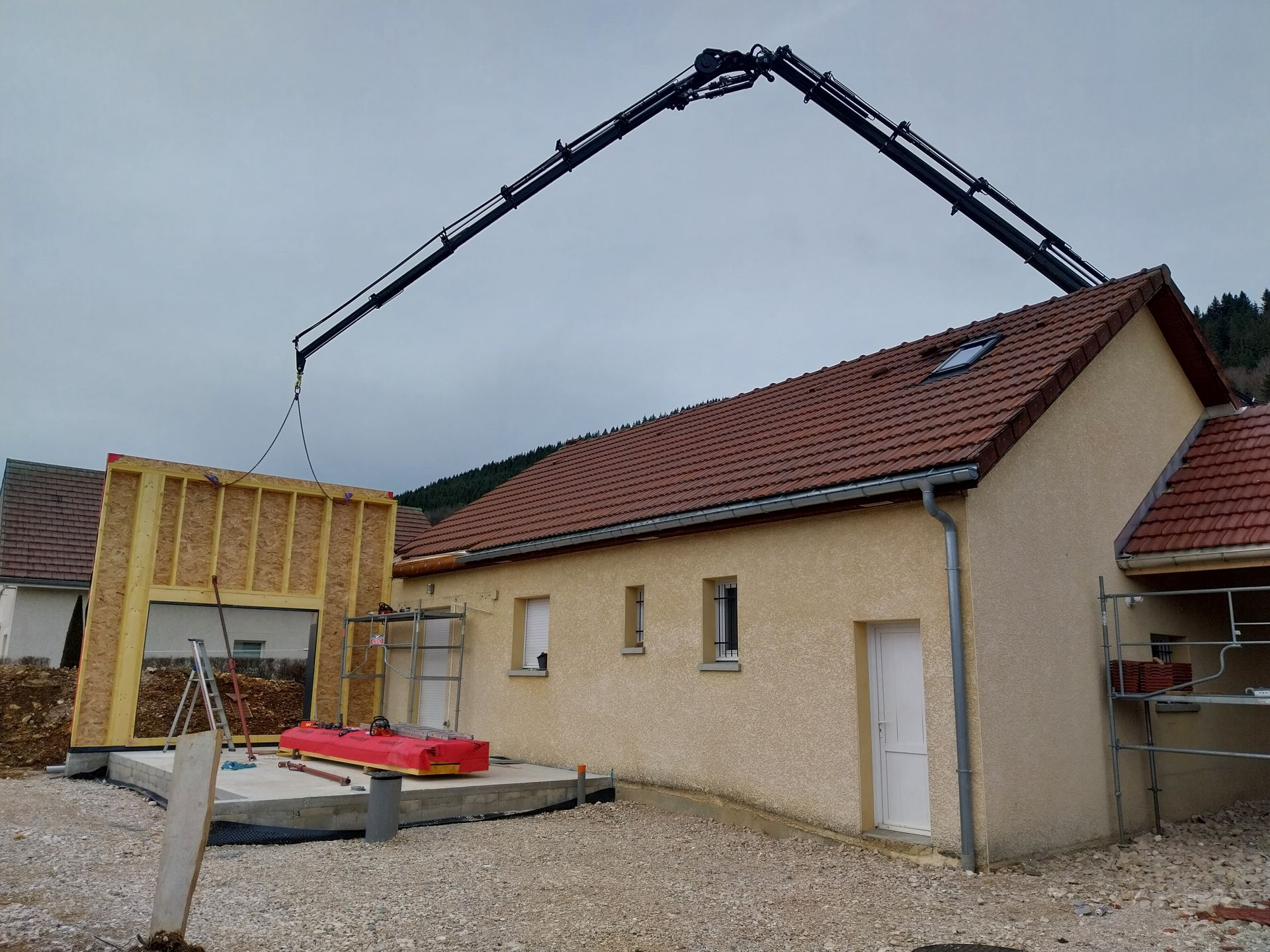 Une grue soulève un panneau mural en bois pour le poser sur un bâtiment en construction ; murs beiges, toit rouge.