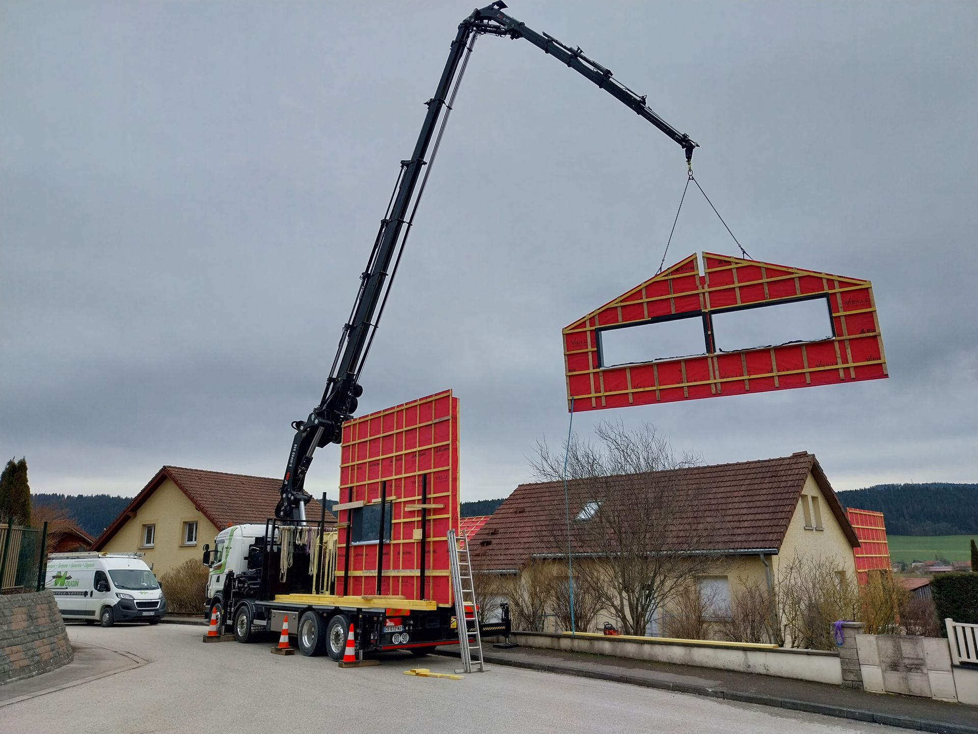 Une grue soulève un pan de mur rouge préfabriqué et le pose sur une maison en construction. Ciel nuageux, quartier résidentiel.