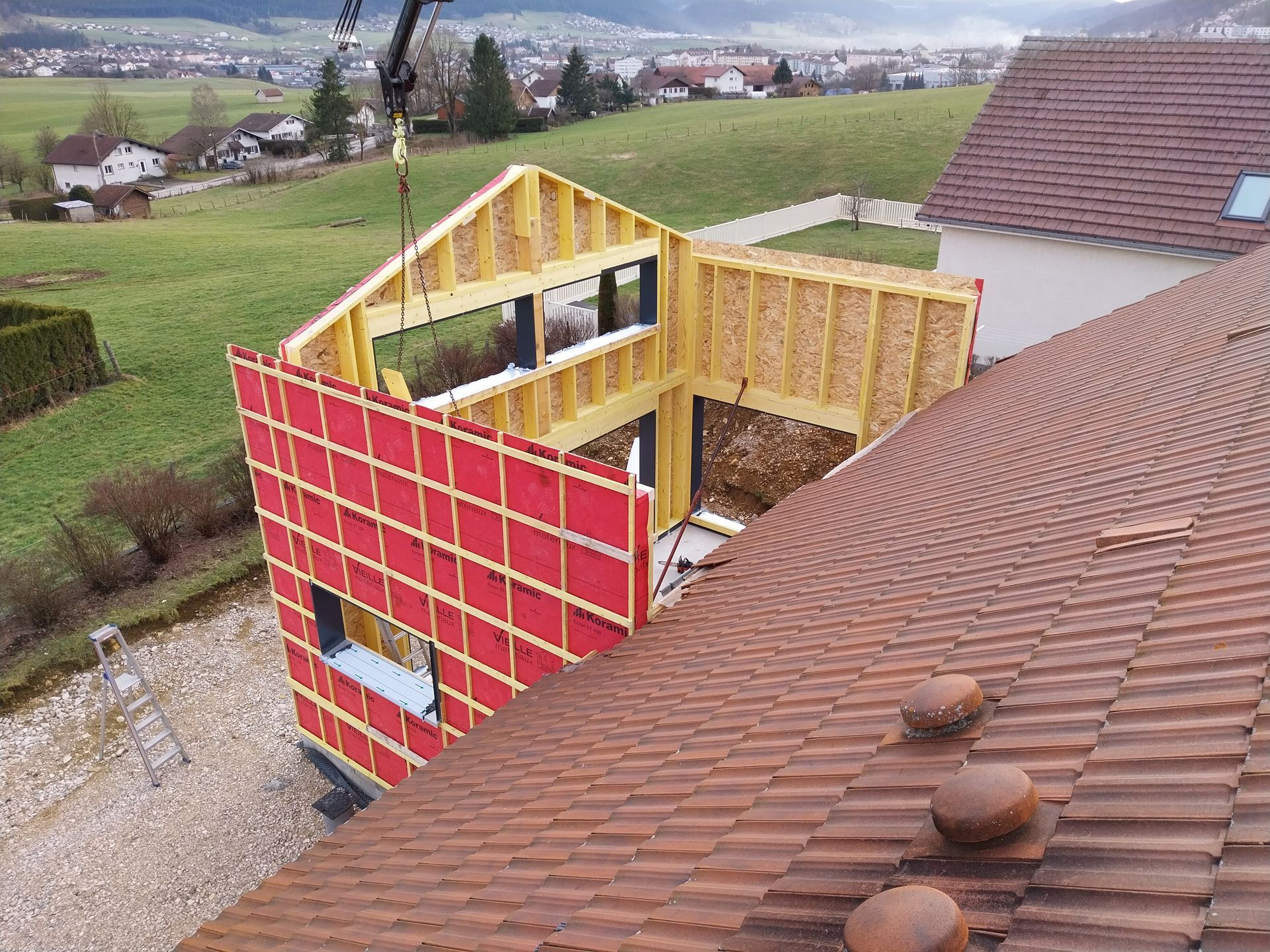 Panneau mural d'une maison soulevé par une grue lors de sa construction, avec un revêtement rouge.