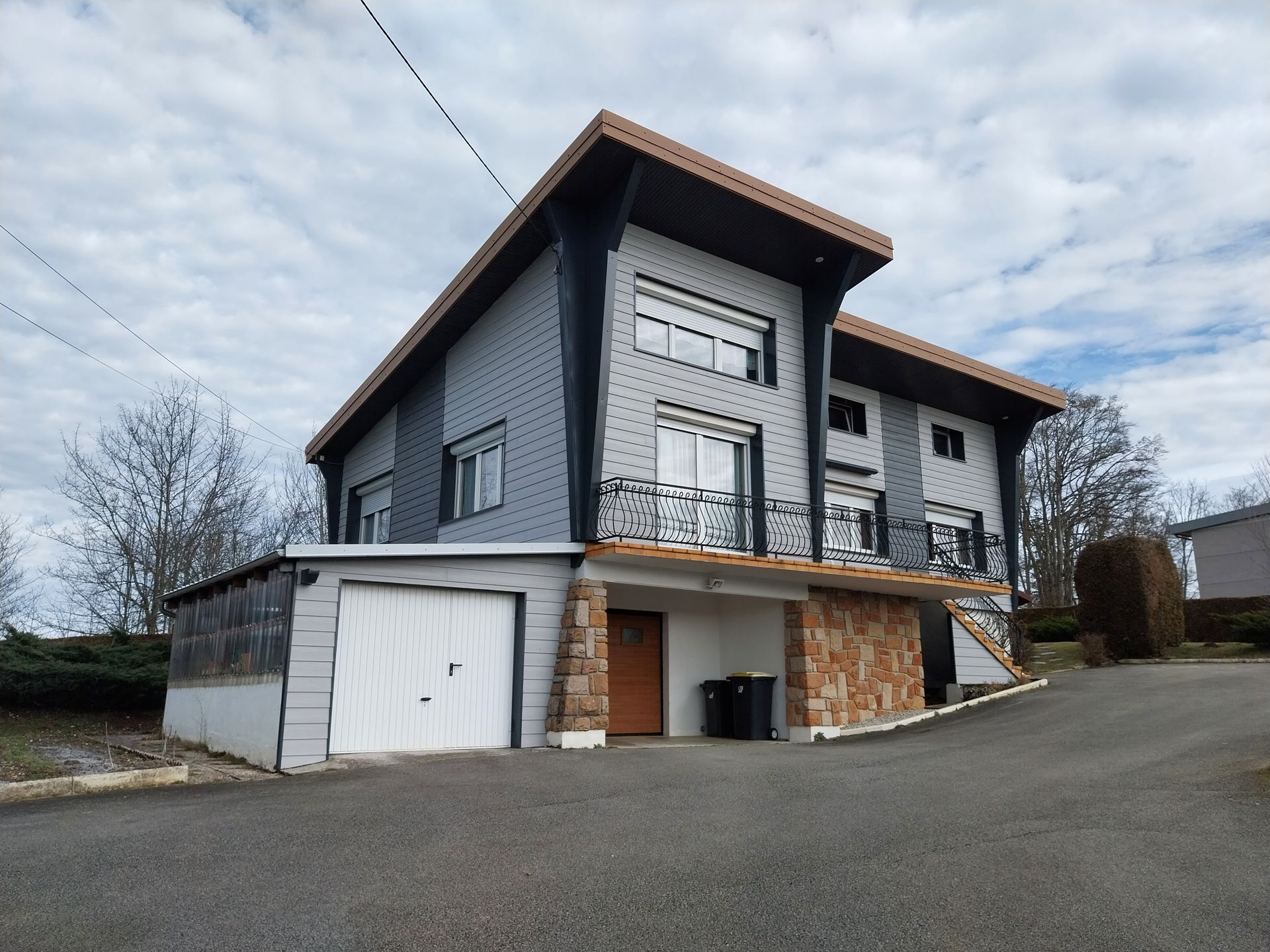Maison moderne à deux étages avec bardage gris, touches de brun, balcons et garage attenant sur une allée en pente.
