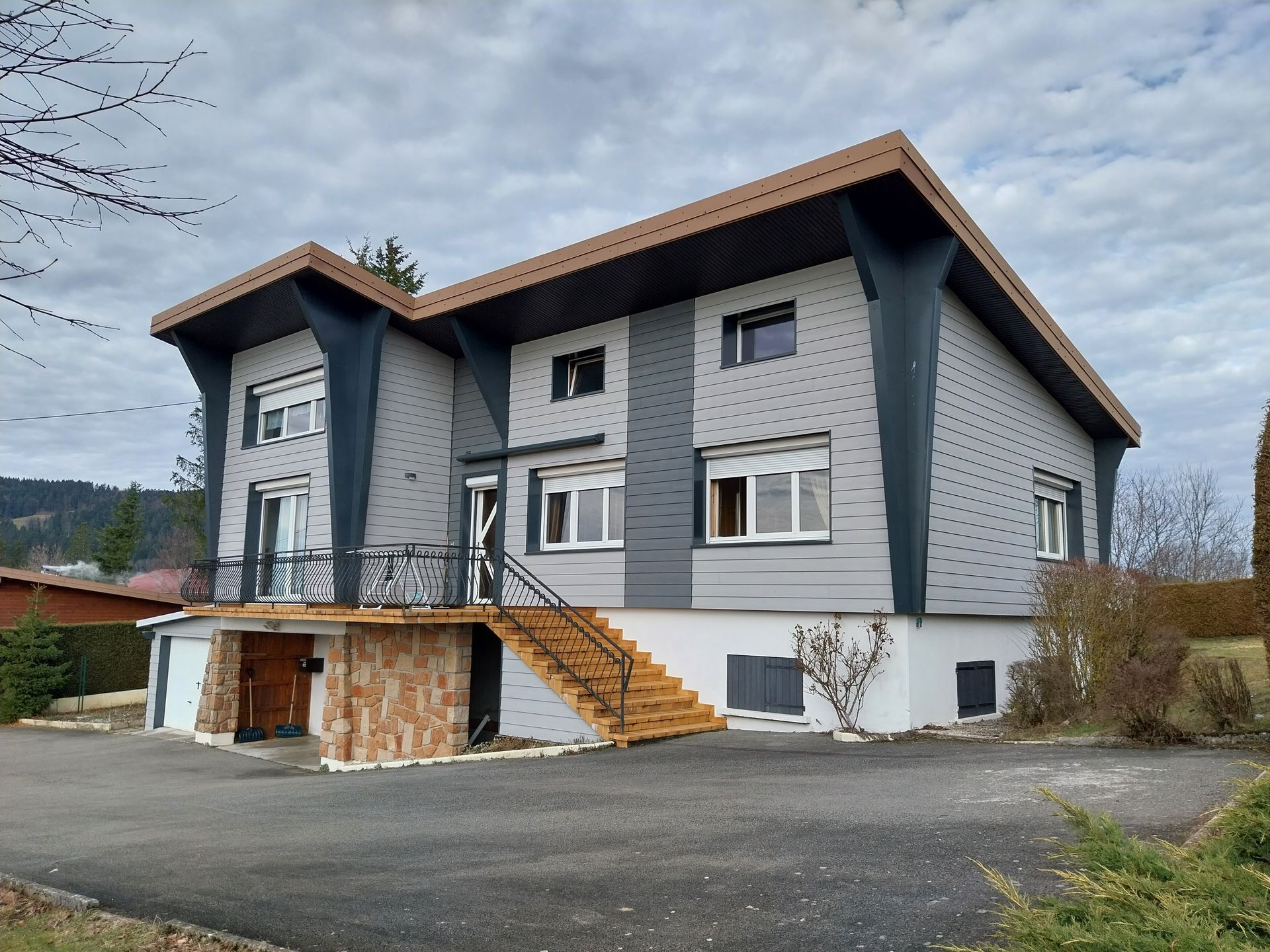 Maison à deux étages avec bardage gris, boiseries foncées, toit marron et escalier en bois menant à la porte d'entrée.