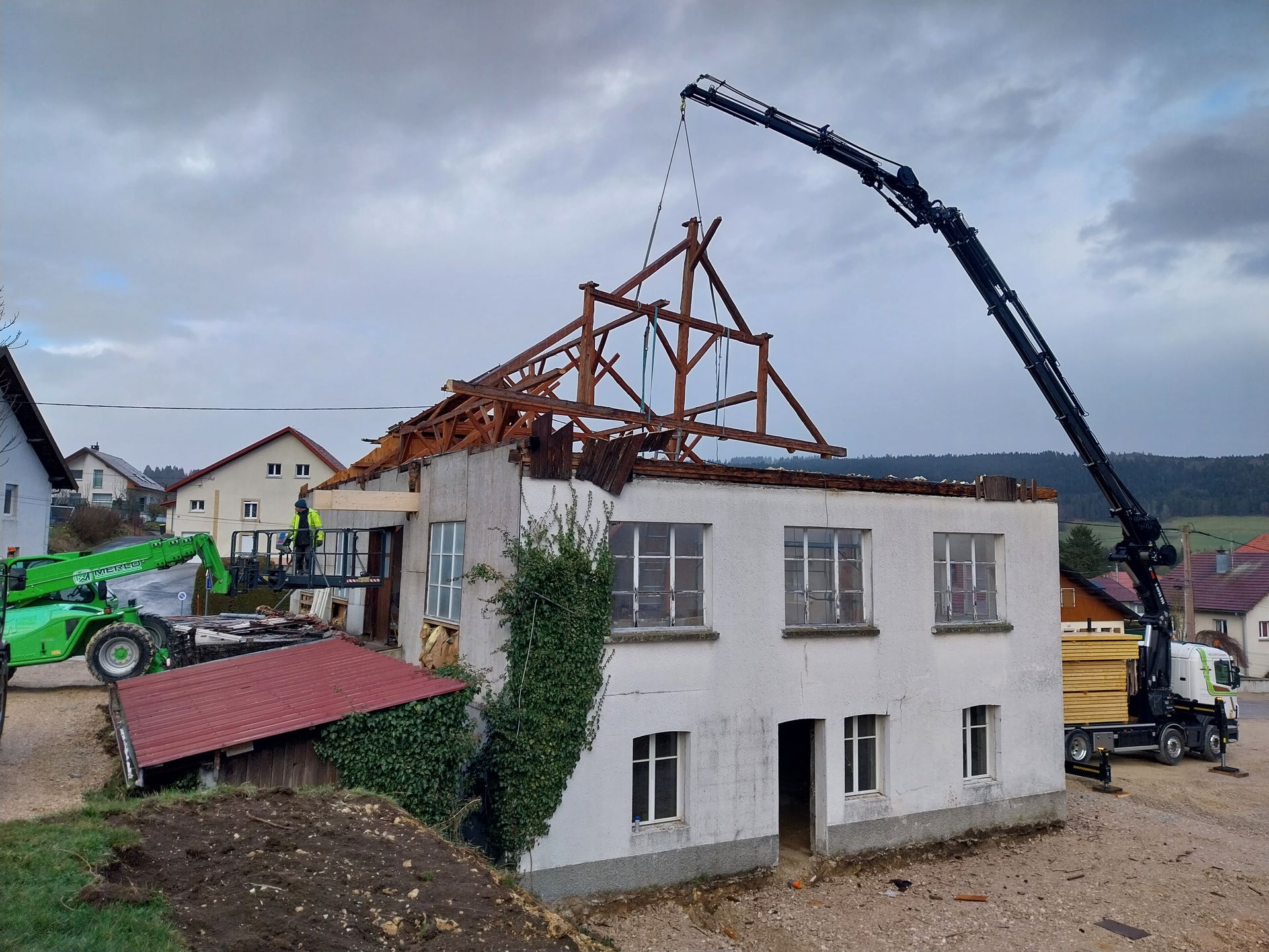 Grue enlevant le toit d'une maison, journée nuageuse, paysage rural. Chariot élévateur vert, camion, bâtiment endommagé.