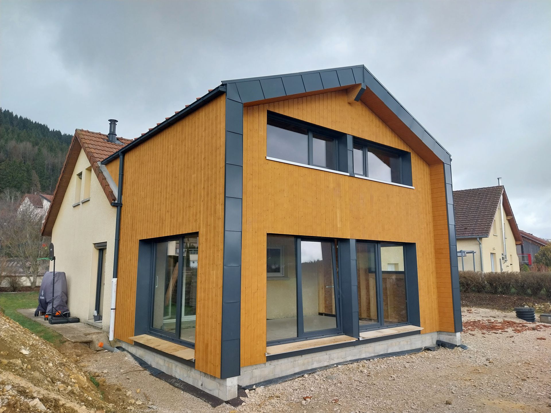 Extension moderne en bois d'une maison, avec de grandes fenêtres et des finitions gris foncé, sous un ciel nuageux.