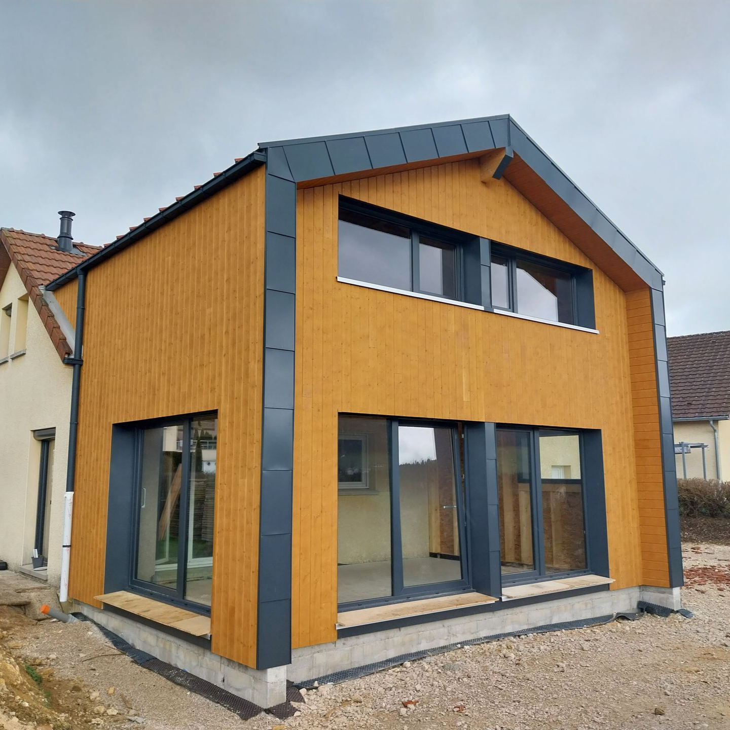 Extension de deux étages avec bardage en bois, boiseries et fenêtres gris foncé, base en gravier.