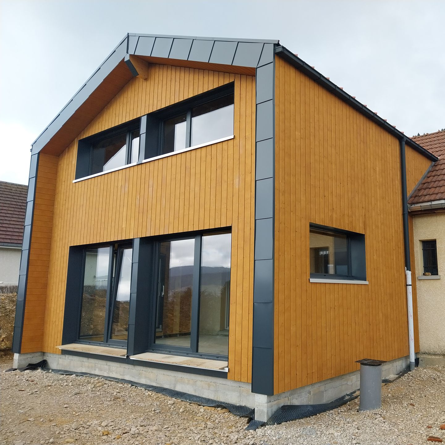 Extension moderne de maison revêtue de bois avec des finitions et des fenêtres foncées ; installée sur des fondations en gravier.