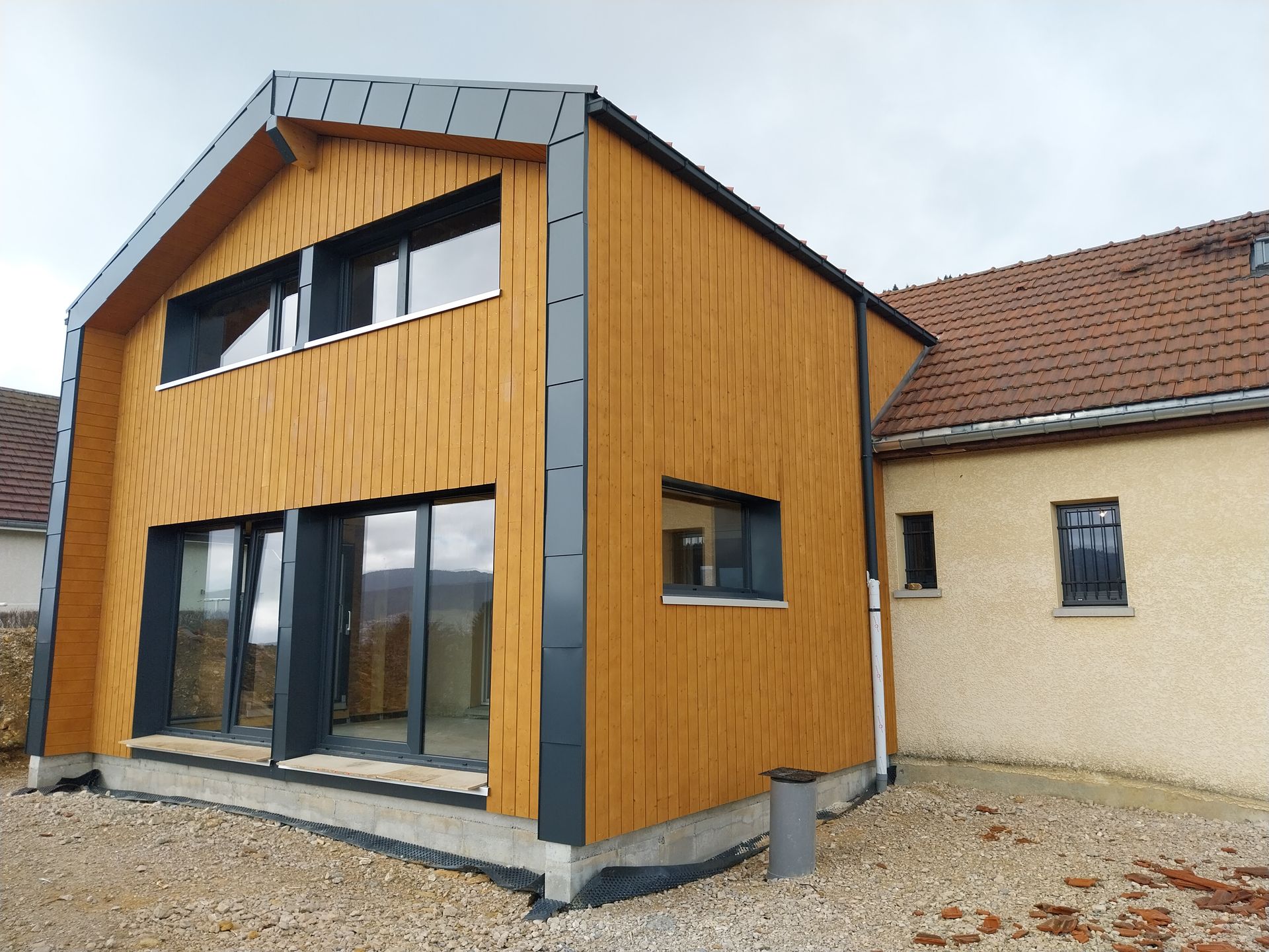 Extension moderne de maison revêtue de bois avec des finitions gris foncé, reliée à un bâtiment plus ancien de couleur beige.