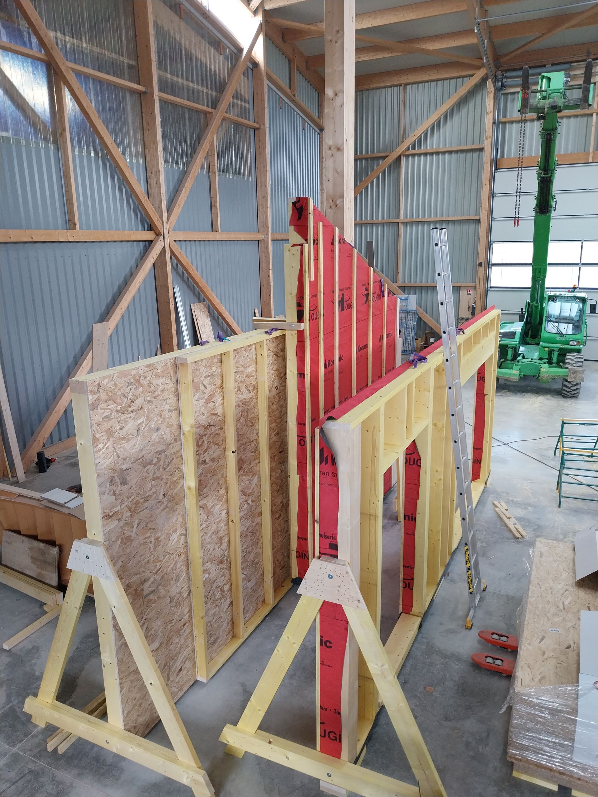 Des murs à ossature en bois sont en construction à l'intérieur d'un entrepôt. Certains murs sont rouges, d'autres sont recouverts de panneaux de bois. Un ascenseur vert est visible.