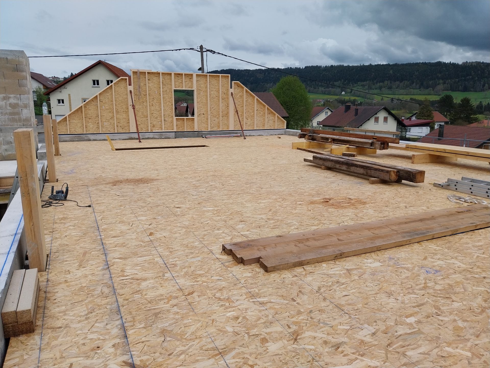 Chantier : toiture en panneaux de bois avec un mur en bois partiellement construit. Journée nuageuse, maisons en arrière-plan.