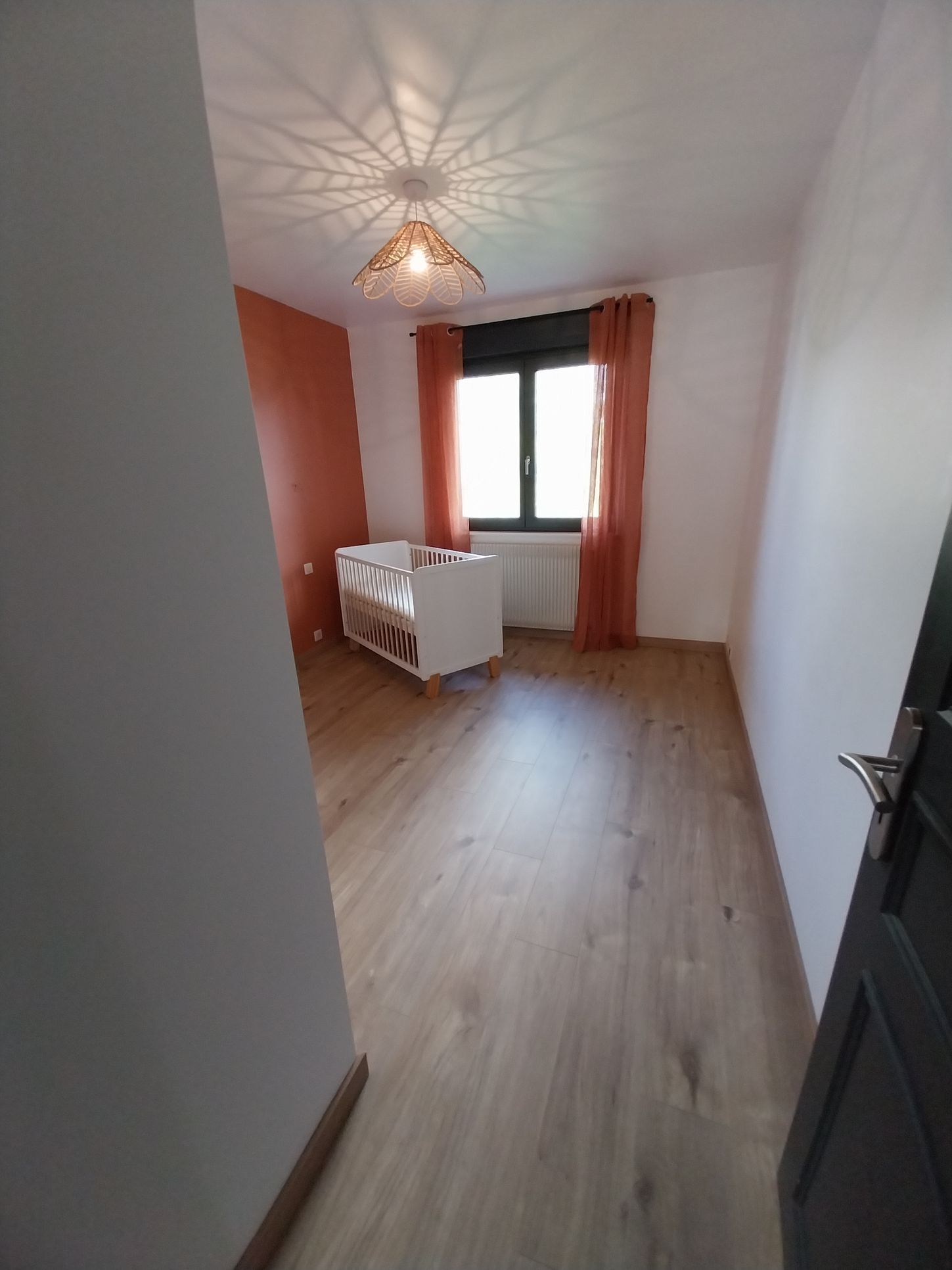 Chambre d'enfant vide avec un berceau, une fenêtre avec des rideaux orange et un sol en bois.
