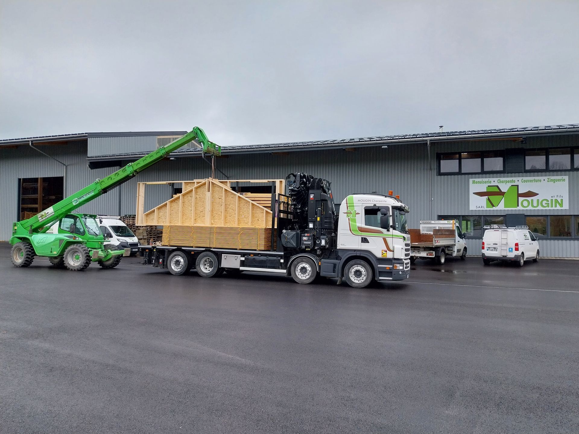 Un chariot élévateur vert charge une structure en bois sur un camion devant un bâtiment.