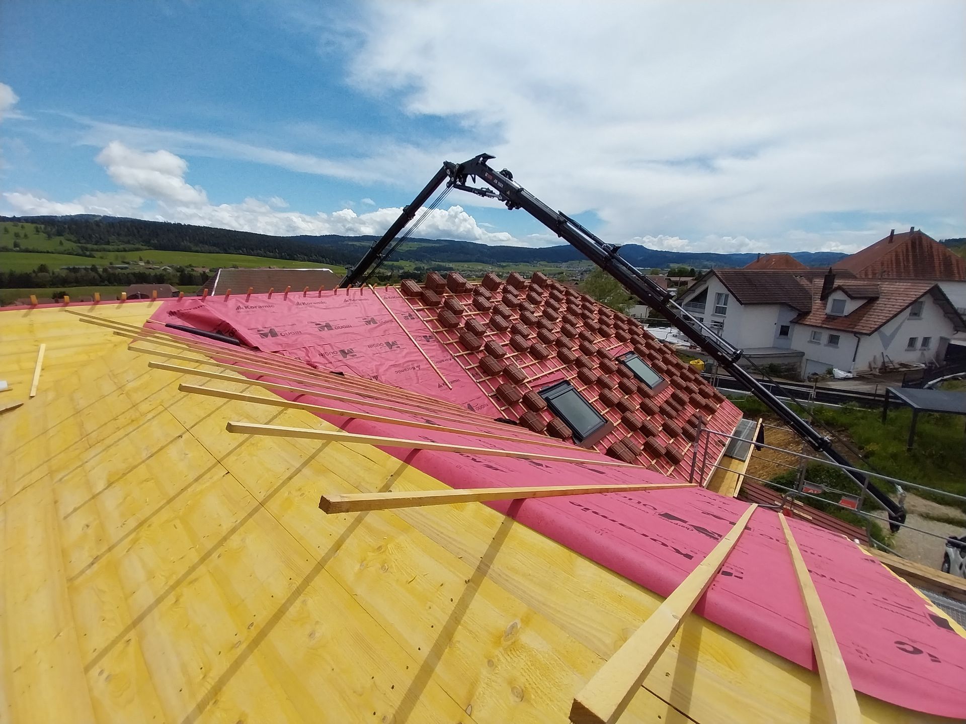 Toiture en construction : isolation rose, tuiles orange, grue, ciel bleu, maisons en arrière-plan.