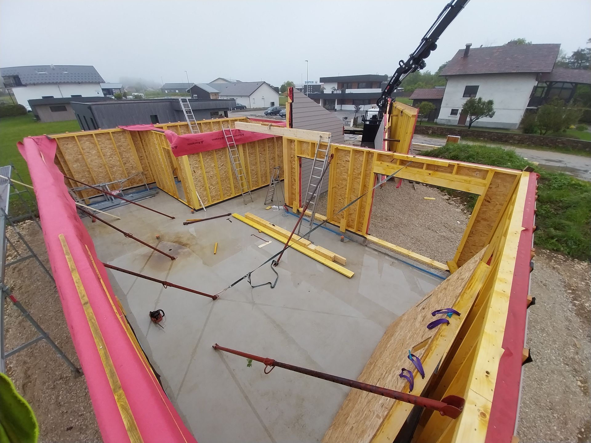 Chantier : maison en bois en construction, grue soulevant des matériaux. Ciel couvert.