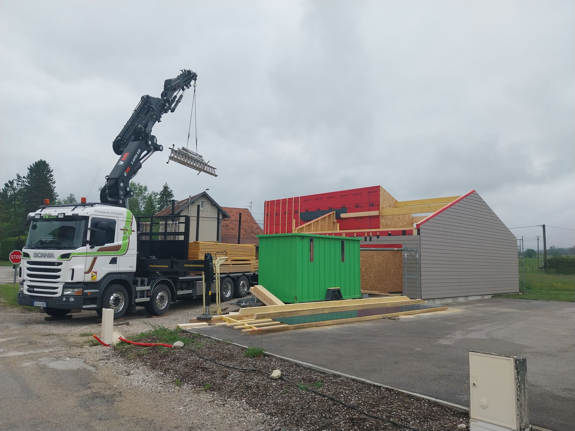 Un camion-grue soulève des éléments de construction en bois sur un chantier sous un ciel nuageux.