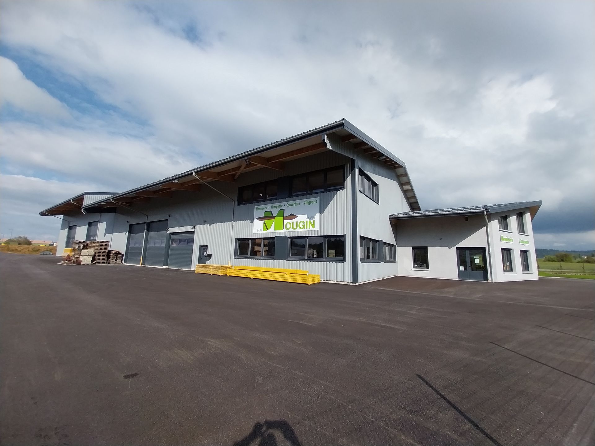 Bâtiment commercial gris au toit en pente. Une enseigne au logo vert est visible. Ciel couvert.