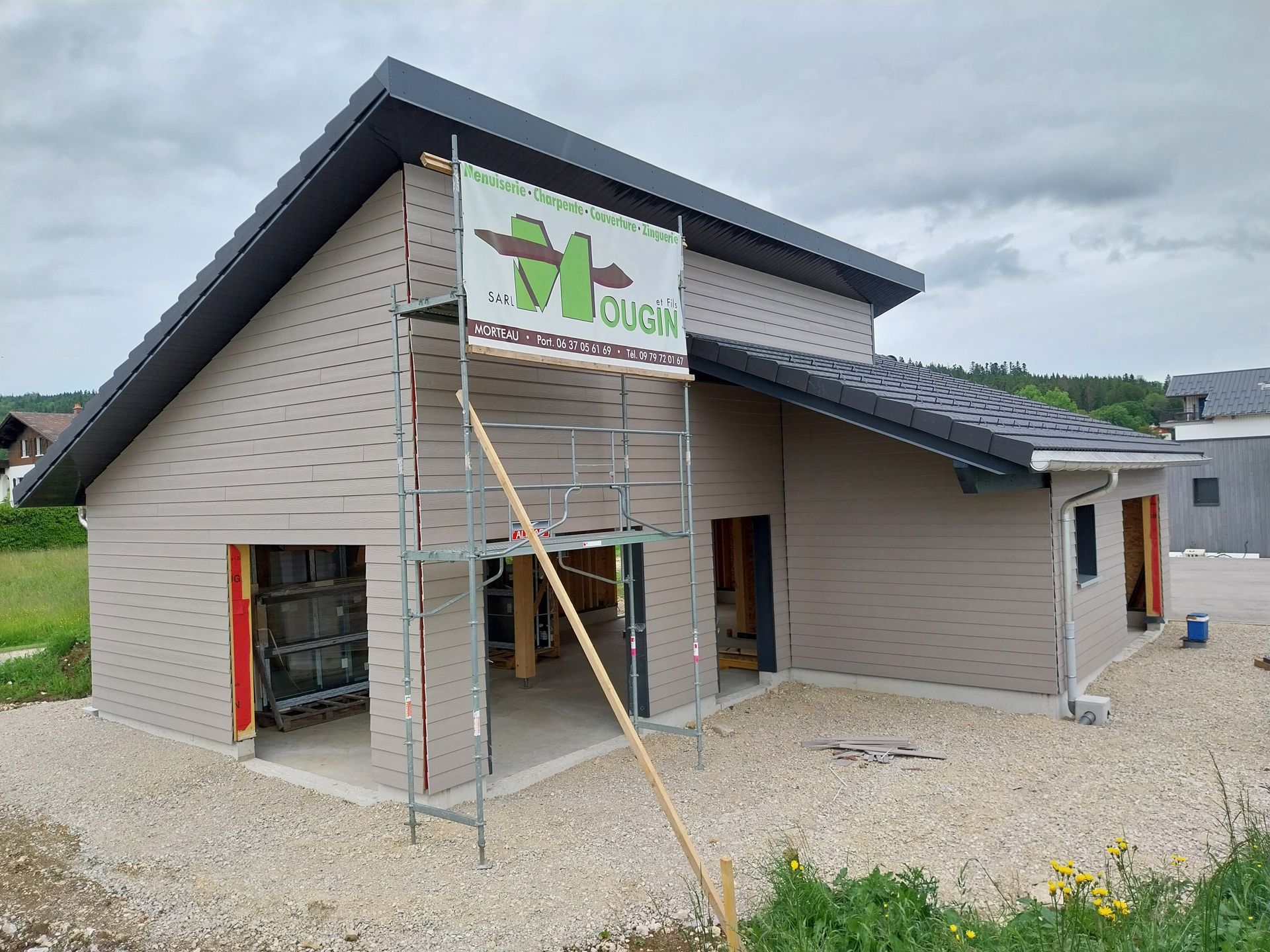 Maison à deux étages en construction avec échafaudages ; bardage gris, toit foncé, cour en gravier, ciel nuageux.