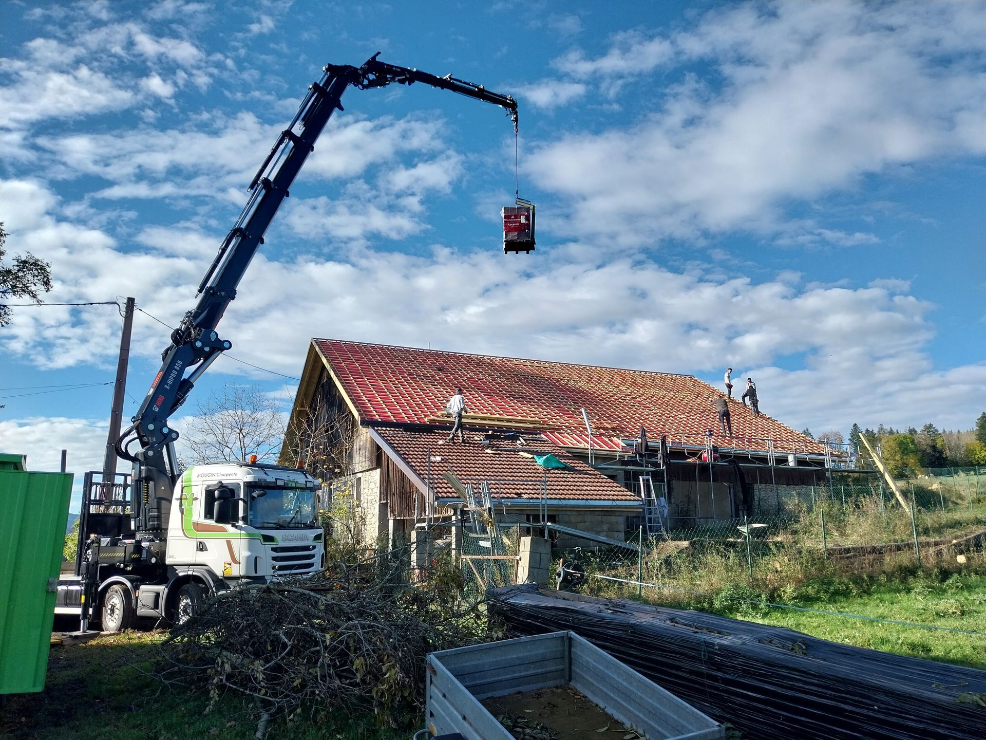 Une grue soulève un conteneur au-dessus d'un bâtiment où des ouvriers se trouvent sur le toit, sous un ciel nuageux.