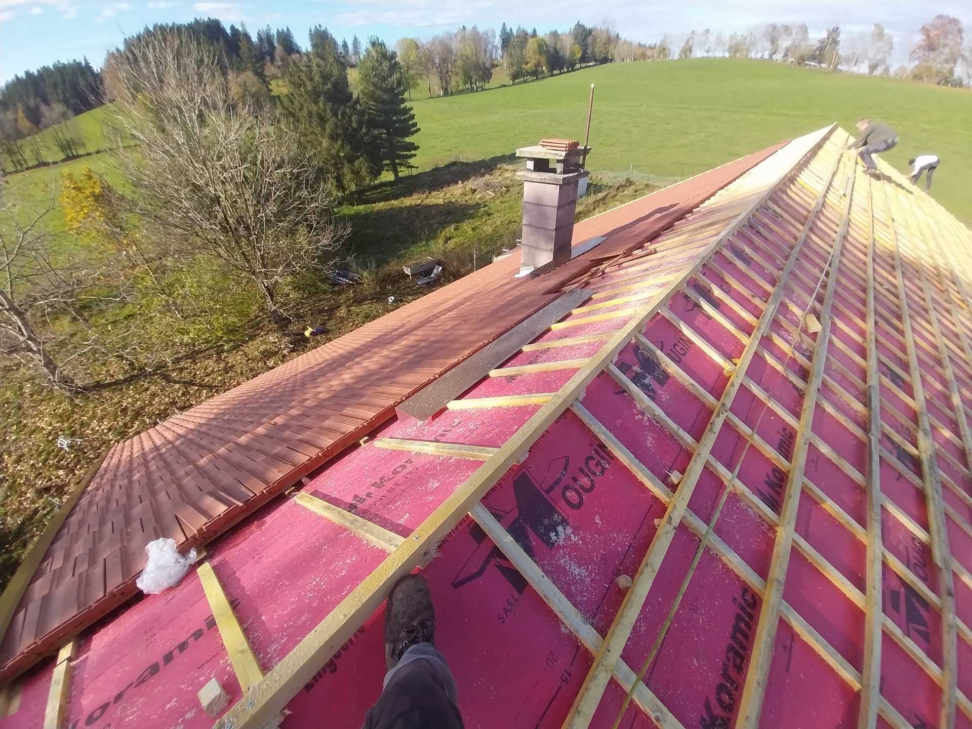 Vue du toit d'une maison aux chevrons apparents, sous-toiture rouge et tuiles en terre cuite. Cheminée et personne sur le toit.