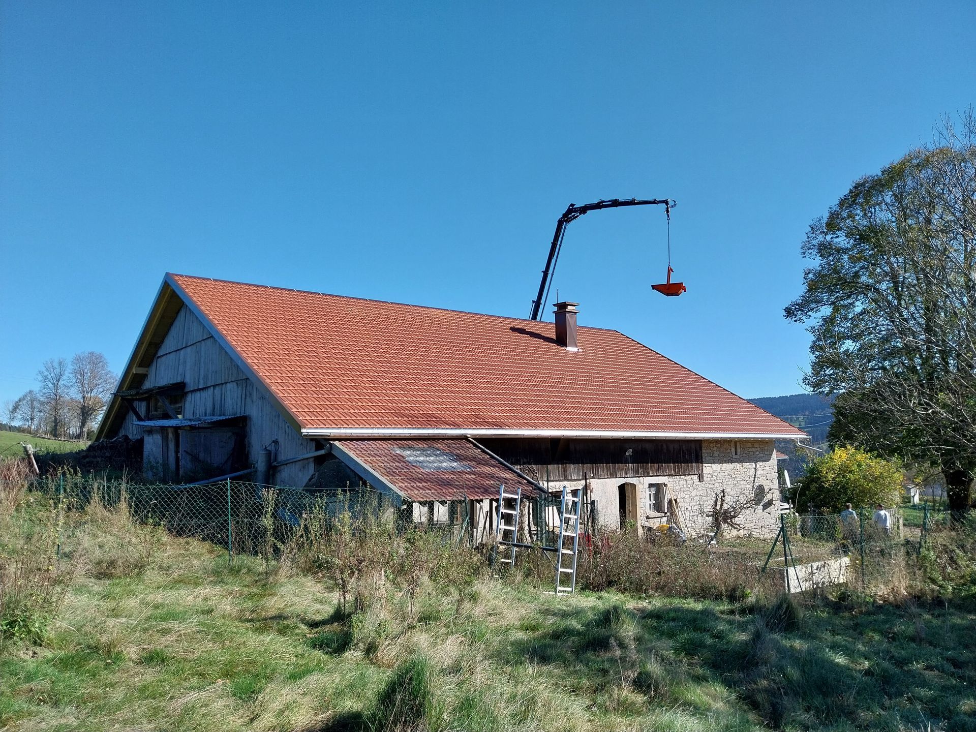 Vieille maison en pierre au toit de tuiles rouges, grue soulevant un objet. Ciel bleu et herbe au premier plan.