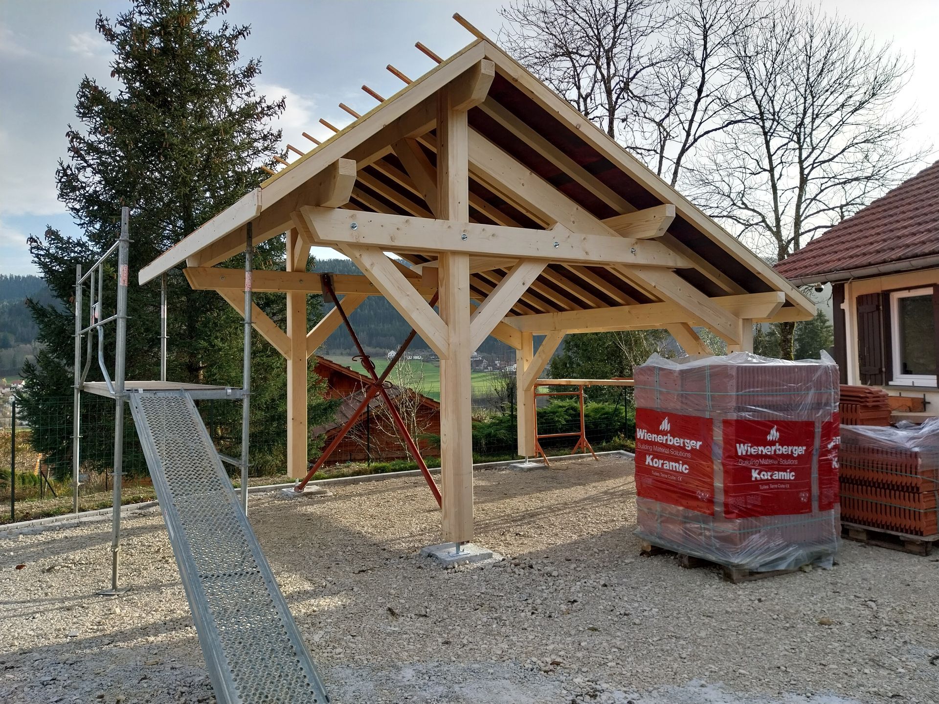 Un abri de voiture en bois est en construction sur du gravier, avec des échafaudages et des briques empilées à proximité.