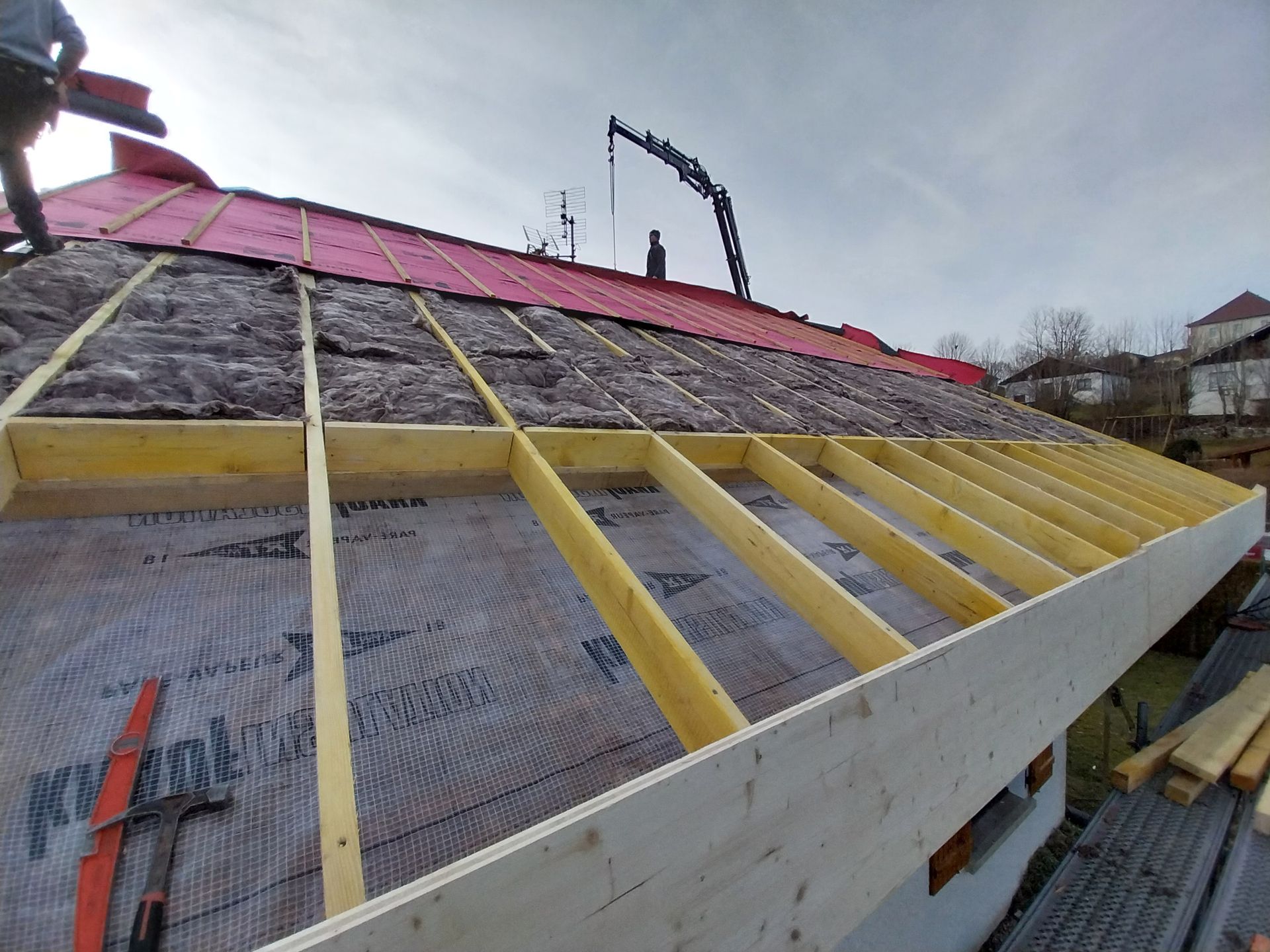 Construction de la toiture : installation de poutres en bois, d'isolant et de matériaux de couverture rouges sur un bâtiment.