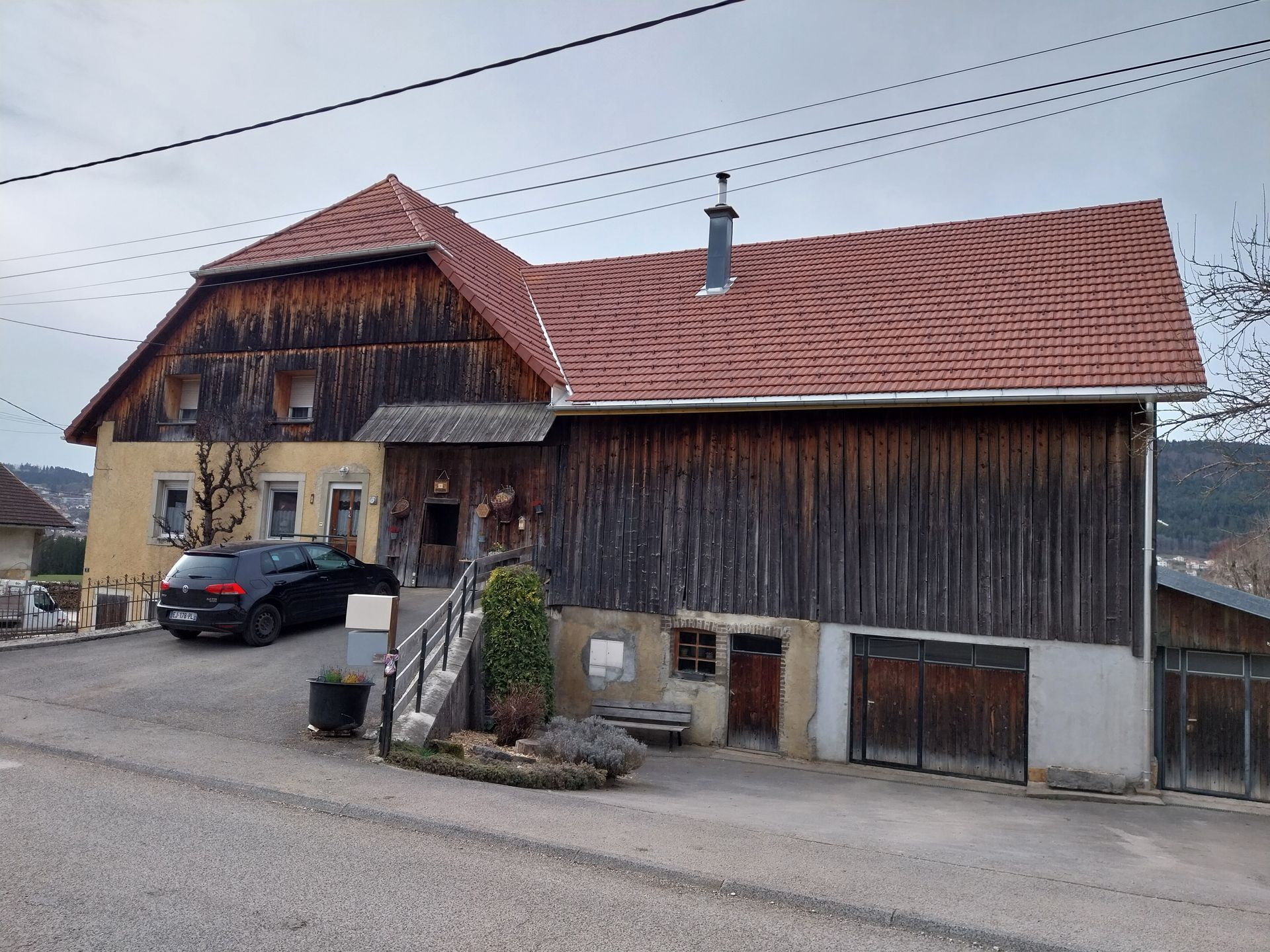 Maison rustique en bois à deux étages, au toit rouge. Une voiture noire est garée devant, dans une rue en pente. Ciel nuageux.