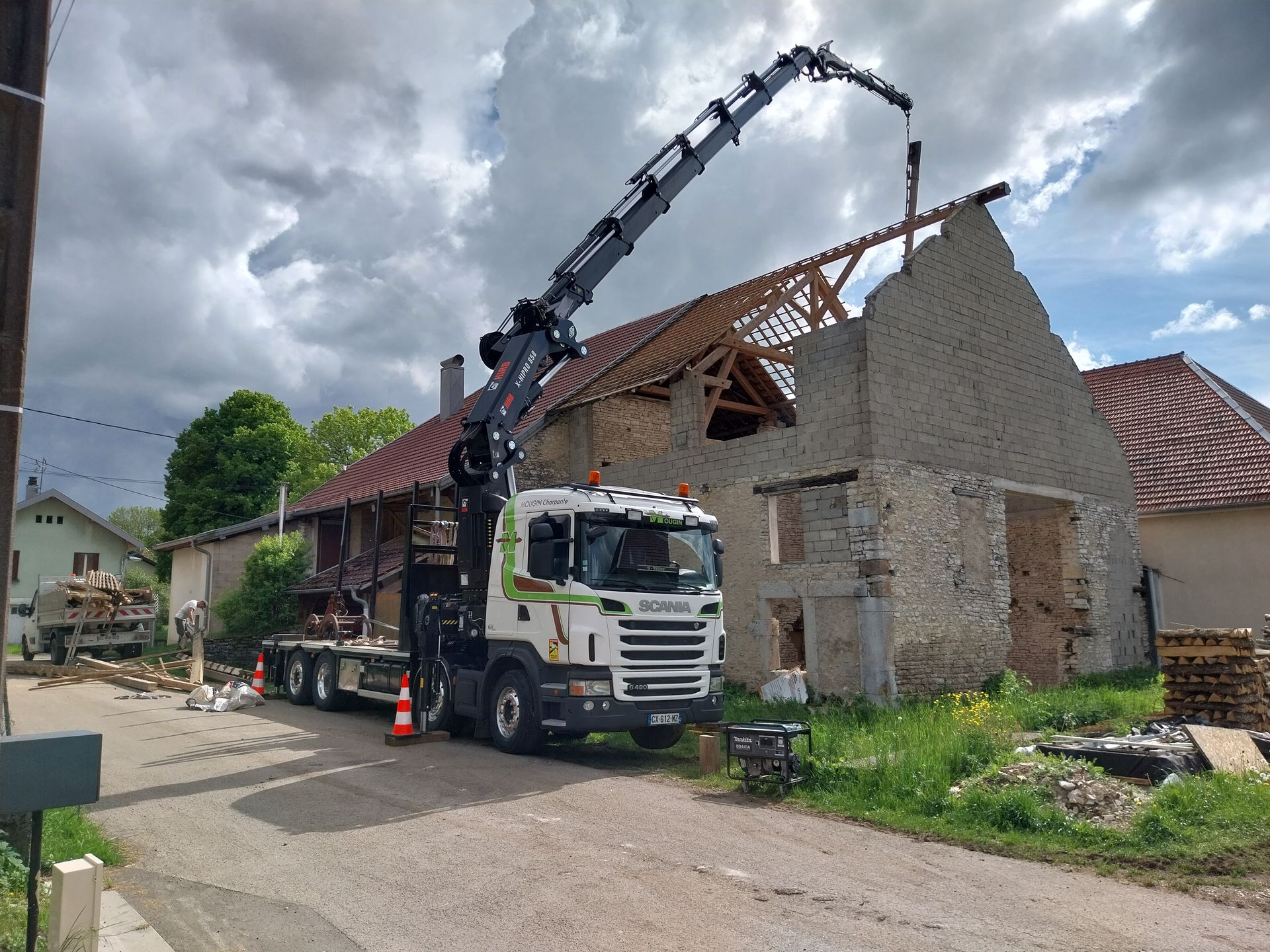 Une grue enlève le toit d'un bâtiment en pierre dans une rue, sous un ciel nuageux.