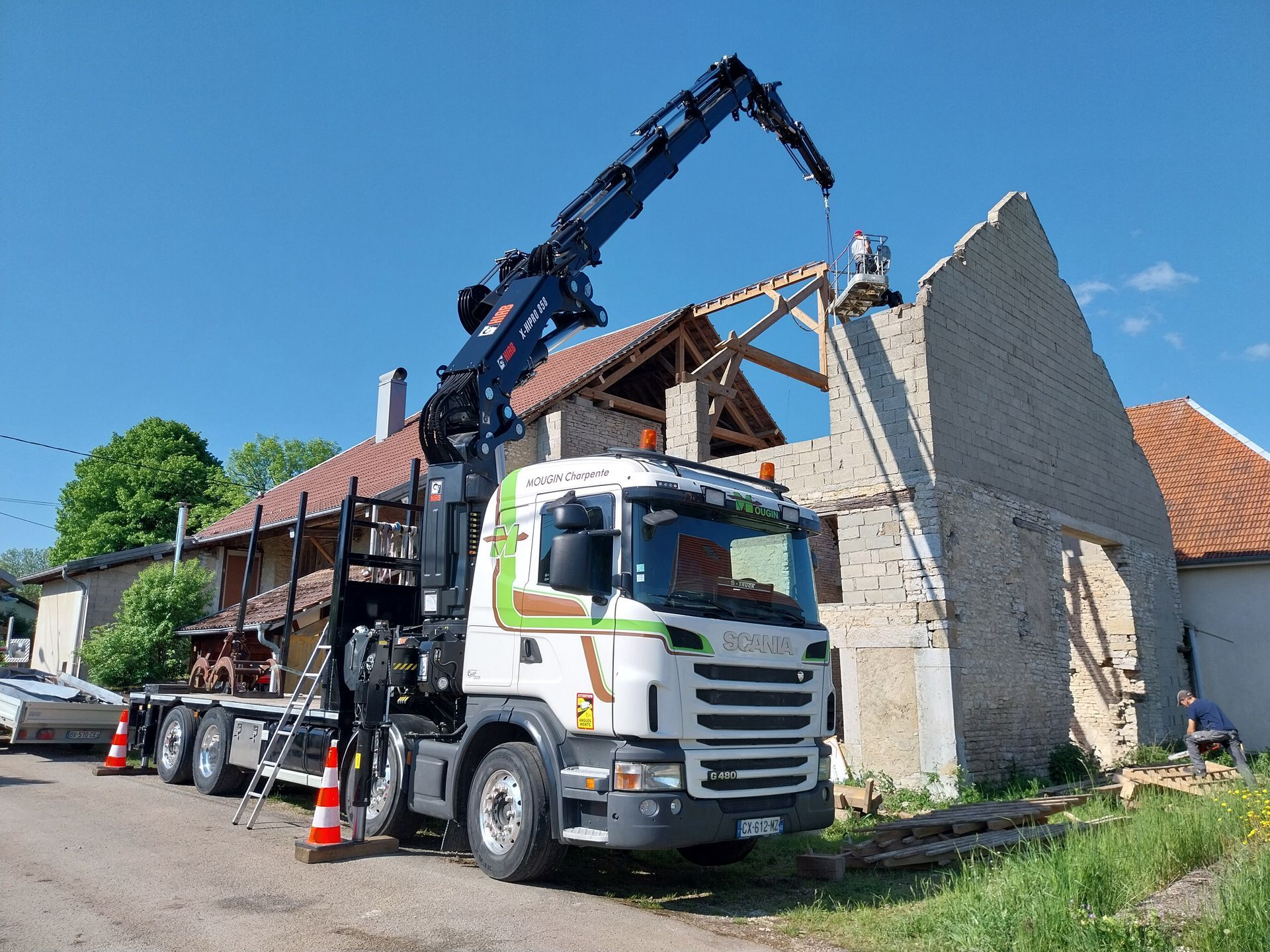 Camion-grue soulevant des poutres en bois pour un bâtiment dont la toiture est partiellement démolie. Journée ensoleillée.