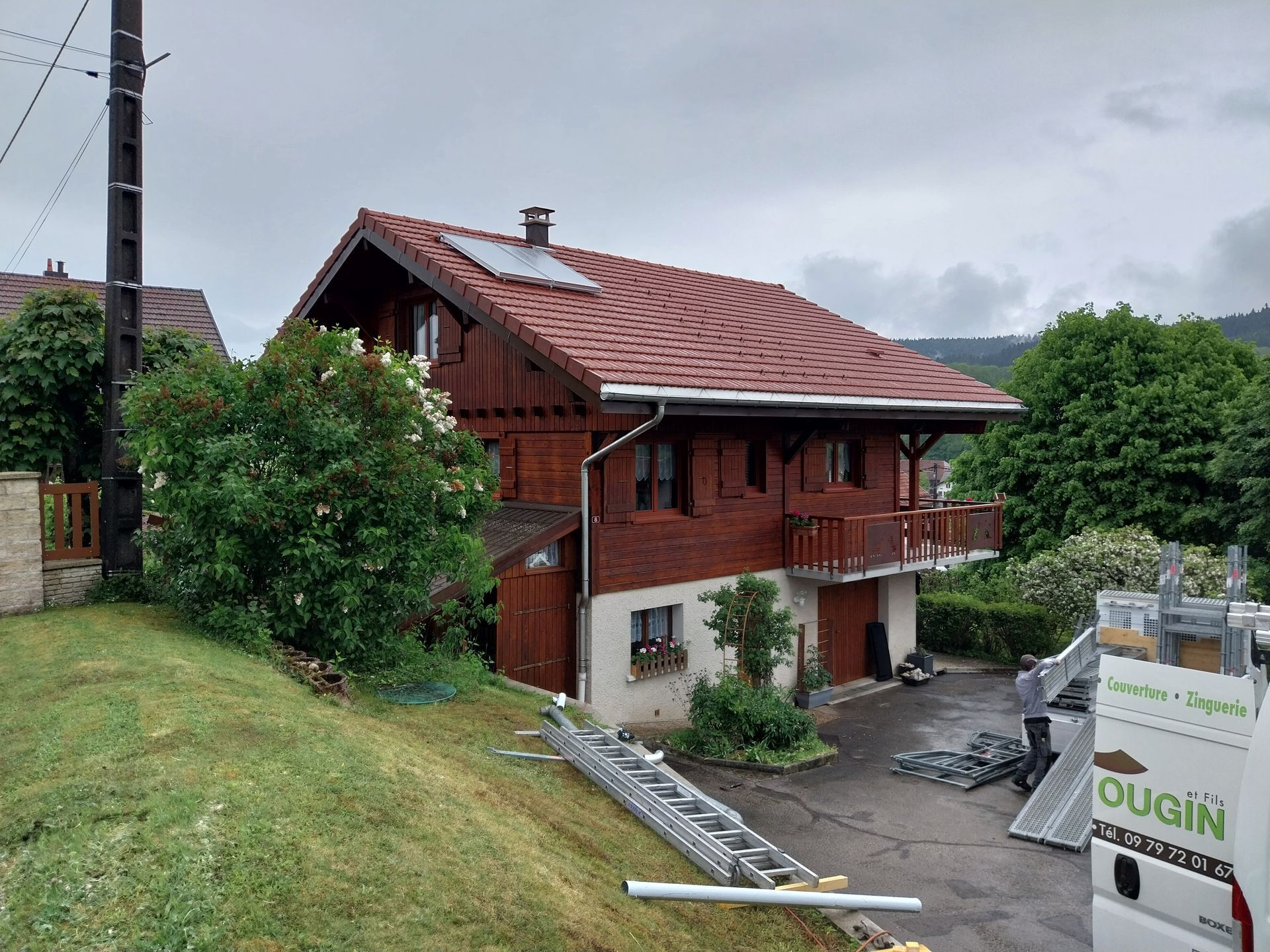Maison en bois au toit brun, avec balcon et garage, située sur une colline herbeuse. Ciel nuageux.