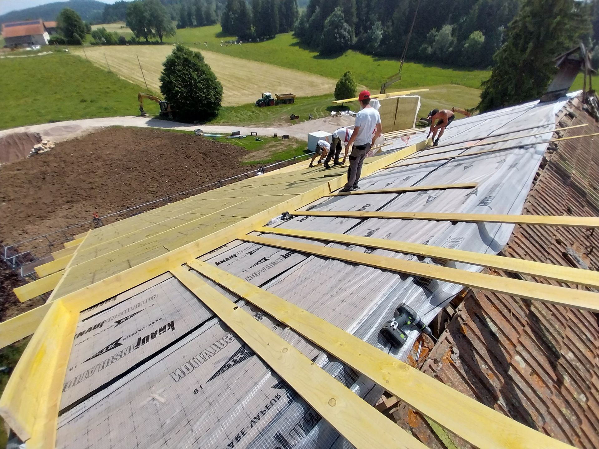 Une équipe de couvreurs installe de l'isolant sur un toit partiellement terminé, avec du bois et un paysage rural en arrière-plan.