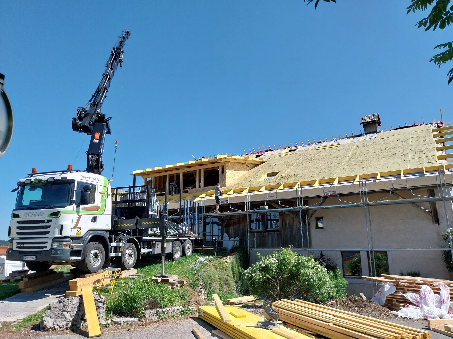 Chantier de construction avec une grue mobile soulevant des matériaux pour la toiture en bois partiellement construite d'un bâtiment. Ciel bleu.