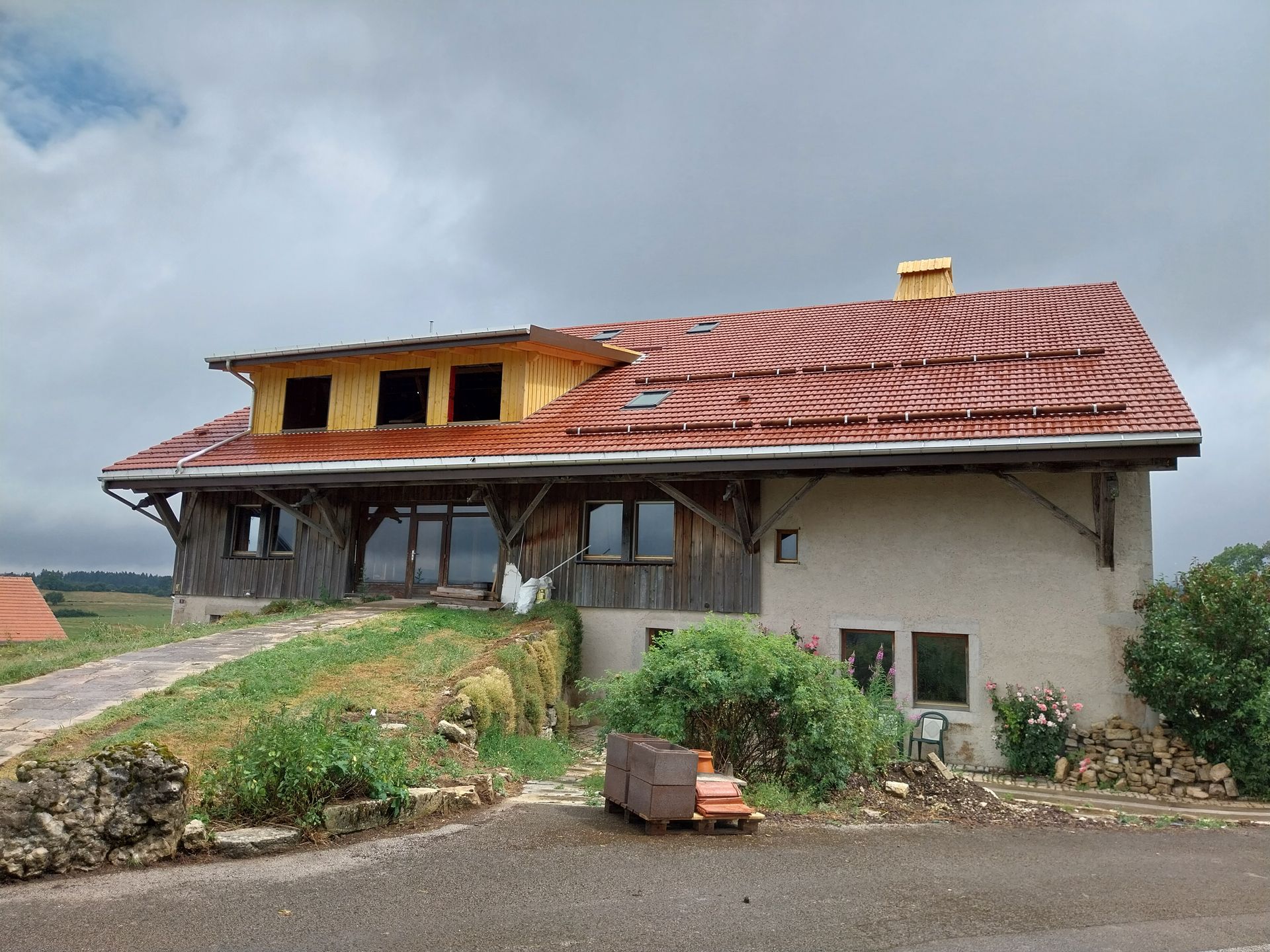 Maison rustique au toit rouge ondulé, ornée de boiseries et de murs en stuc, se détachant sur un ciel nuageux.