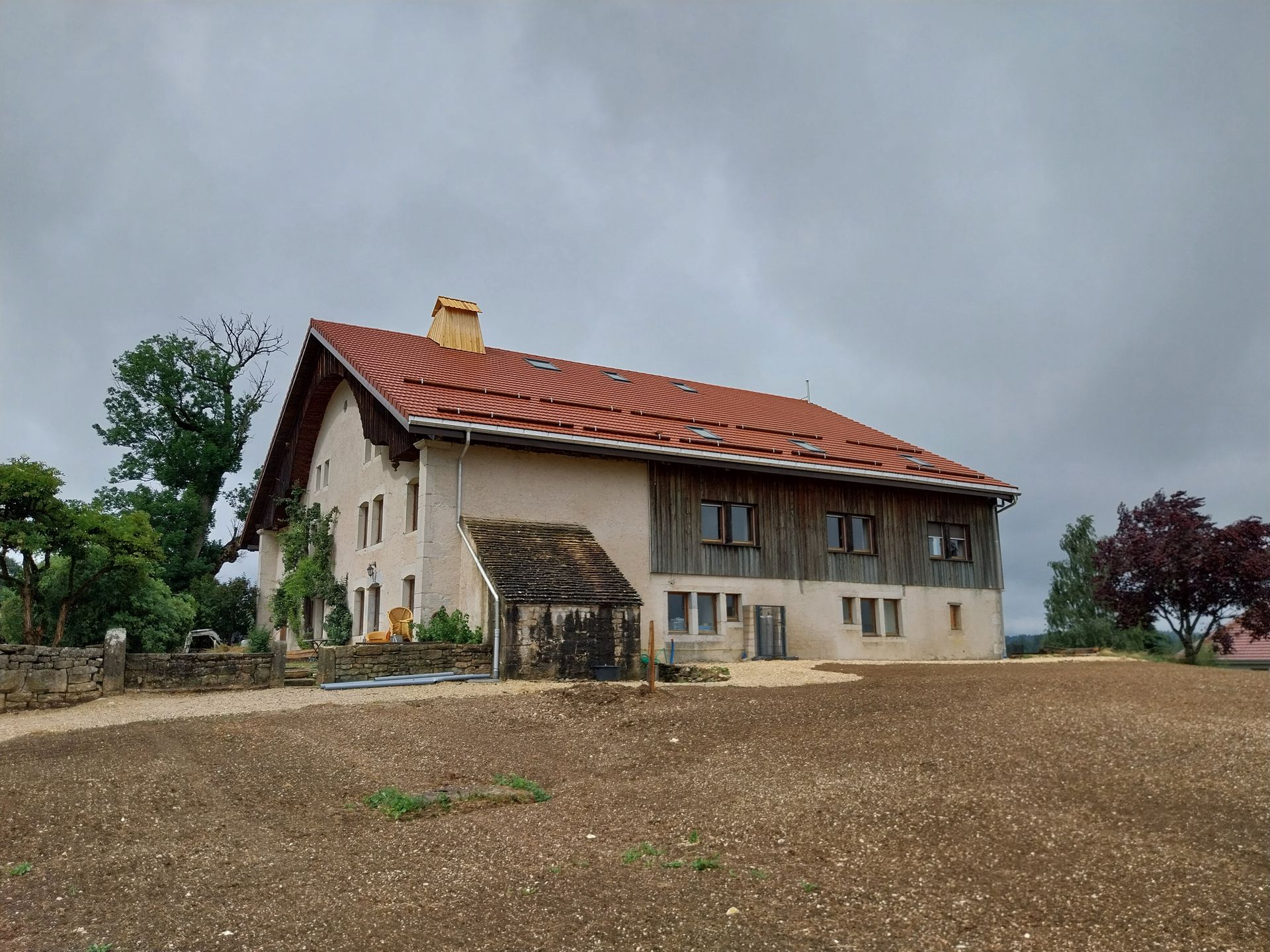 Ferme à deux étages avec toit de tuiles rouges et bardage en bois, sous un ciel nuageux.