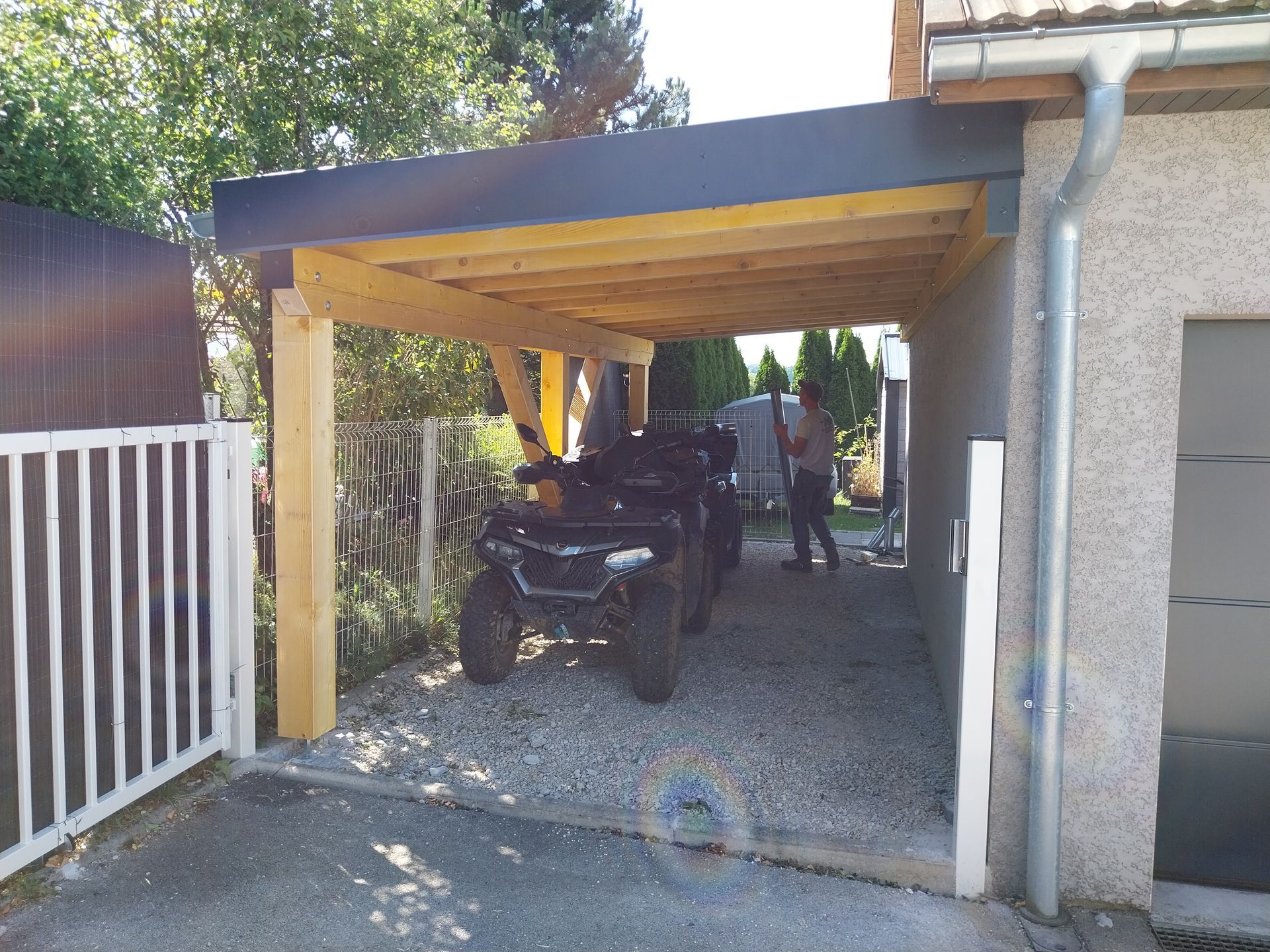 Un quad est abrité sous un abri en bois. Une personne se tient à proximité. Sol gravillonné.